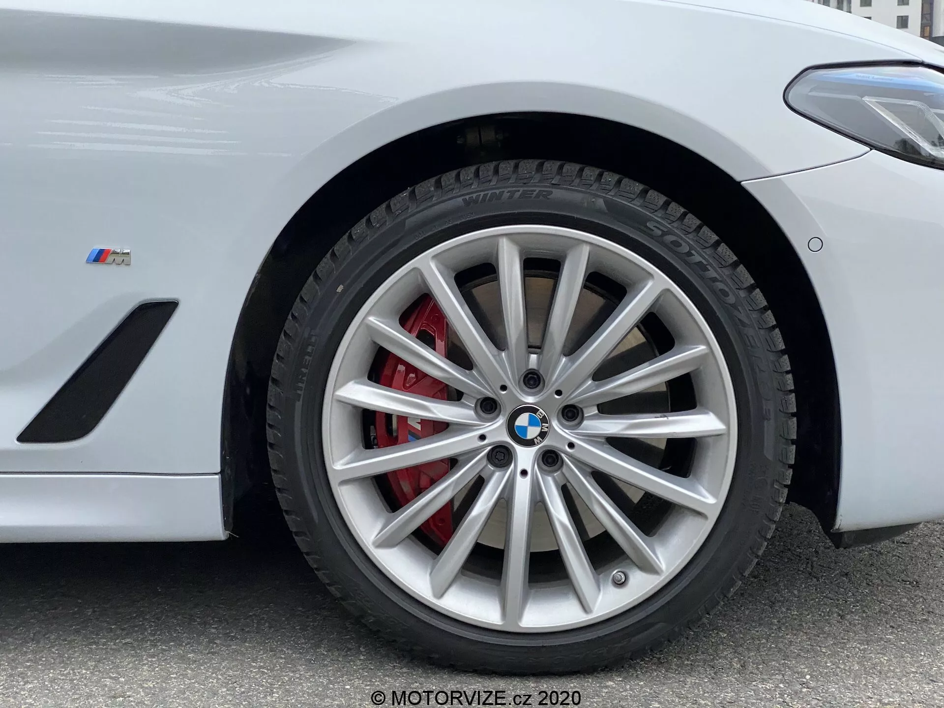 Vue en gros plan de la section de la roue avant d'une BMW de la Série 5 de 2020, spécifiquement le modèle 520i Touring Steptronic. La photographie en contre-plongée met en avant le design complexe de la jante en alliage à multiples branches avec une finition argentée, complété par un étrier de frein rouge et un rotor ventilé. L'emblème BMW et le badge 'M' sur l'aile indiquent qu'il s'agit d'un véhicule de performance de la série M de BMW. L'image capture des détails clés adaptés pour présenter des systèmes de freinage haute performance, des éléments de design de véhicules de luxe et des technologies de pneus.