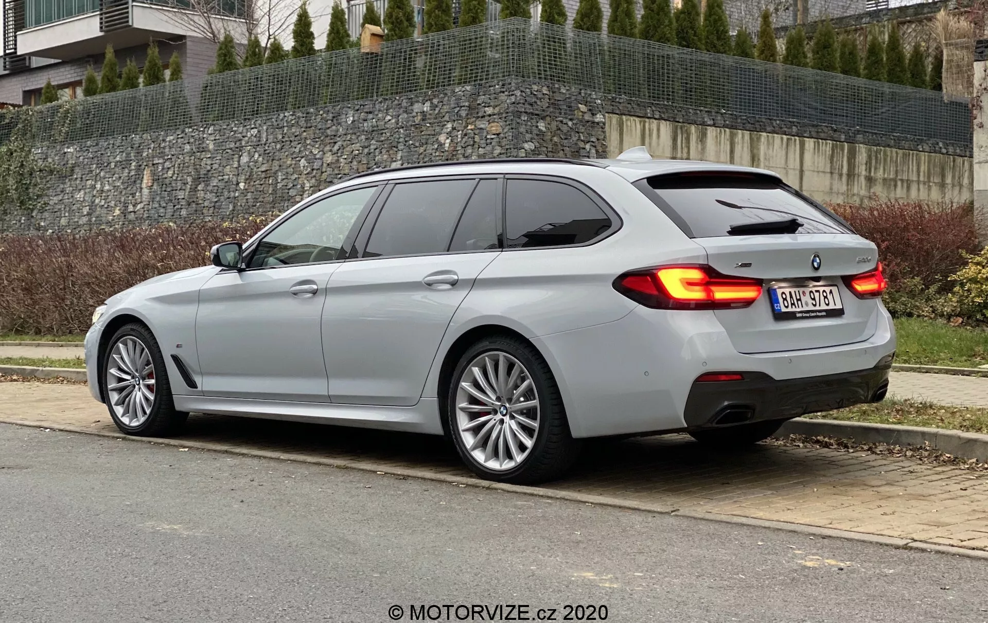 L'image montre une voiture break moderne vue sous un angle arrière trois quarts, mettant en valeur la ligne de toit élégante et le profil latéral du véhicule. Elle met en évidence les feux arrière horizontaux s'étendant légèrement dans le hayon, une lunette arrière fortement inclinée pour un look plus sportif, et des accents chromés autour des fenêtres latérales. La voiture a une finition argent métallisé qui reflète la lumière couverte, des roues à rayons multiples indicatives d'un segment de luxe et des doubles tuyaux d'échappement suggérant une variante de moteur plus puissante ou un pack sport. La présence de feux arrière à LED et d'un spoiler arrière subtil ajoute à son esthétique technologique et sportive.