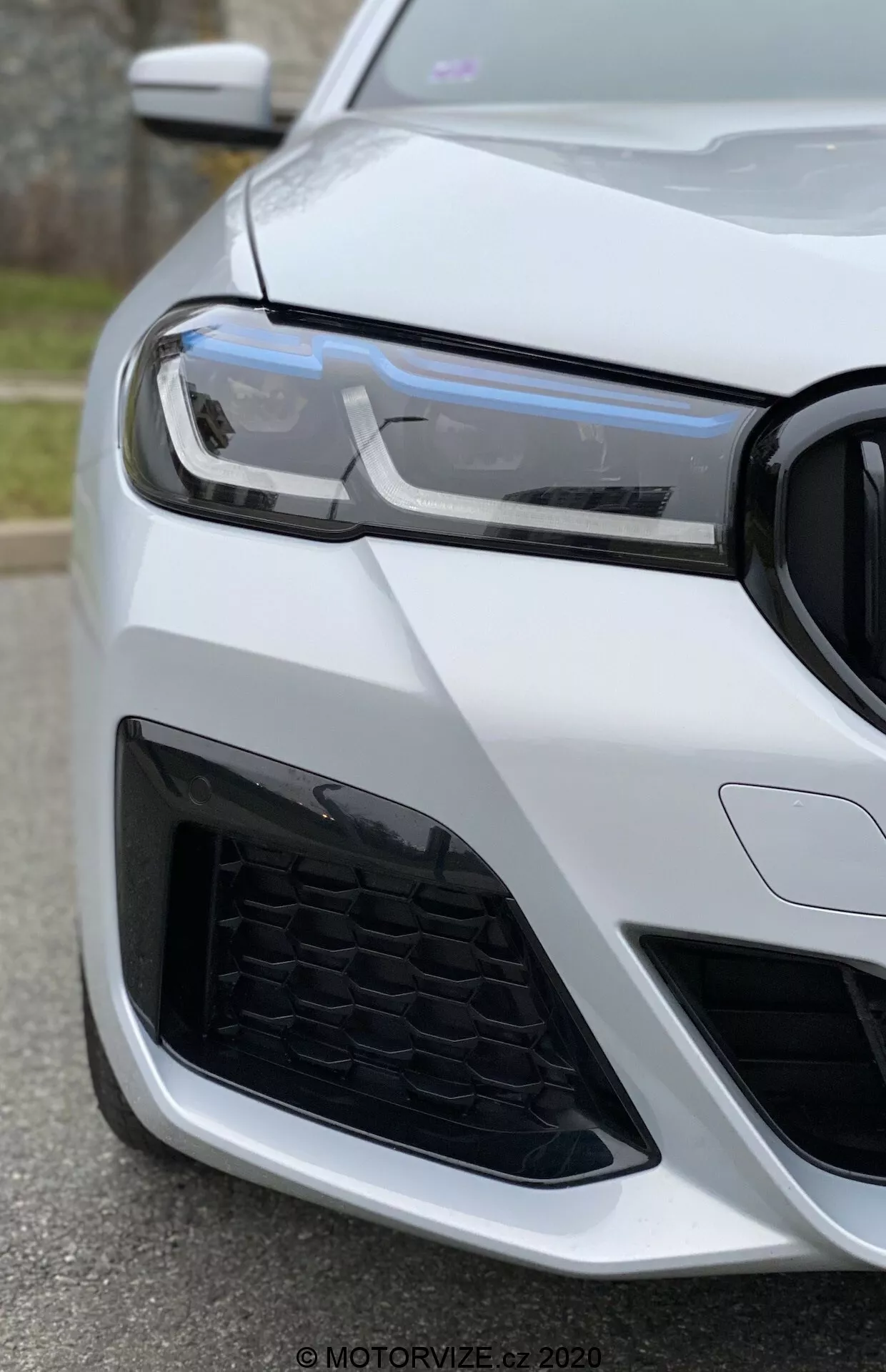 Vue rapprochée du coin avant gauche d'une BMW 520i Touring Steptronic (2020) prise sous un angle bas, se concentrant sur le phare, la calandre et le pare-chocs. Les feux de jour LED modernes, le motif en nid d'abeille de la grille et le design agressif de la prise d'air suggèrent un véhicule sportif et avancé. La voiture est peinte en blanc brillant, accentuant son apparence luxueuse et bien entretenue.