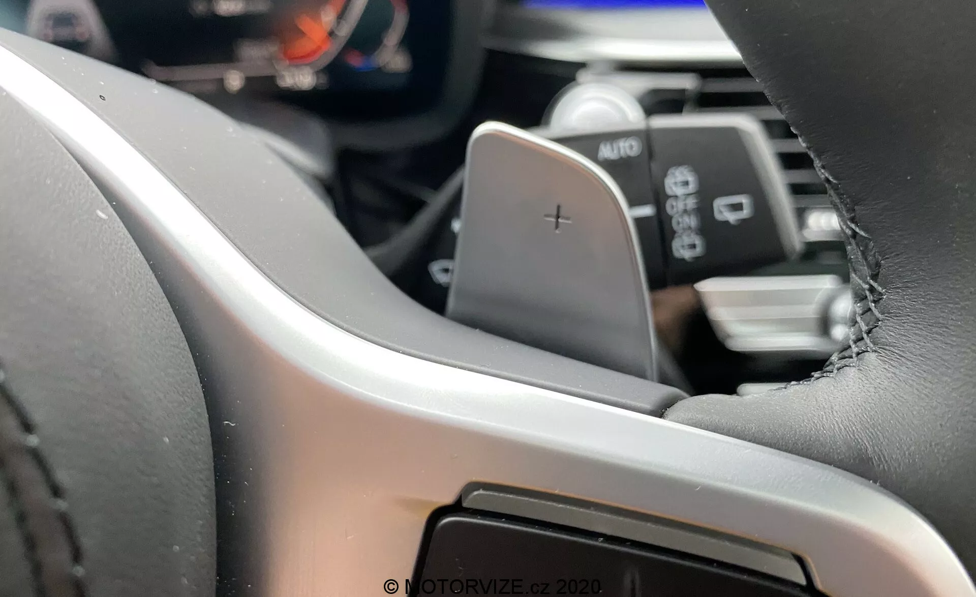 Vue intérieure de la BMW Série 5 2020 (BMW 520i Touring Steptronic) se concentrant sur le côté gauche du volant du point de vue du conducteur⁠—volant moderne en cuir avec coutures visibles, équipé d'une palette de changement de vitesse marquée d'un '+'. Le tableau de bord numérique ou le panneau d'instruments est partiellement visible, mettant en évidence des systèmes d'information avancés pour le conducteur. Sous la palette, une manette marquée AUTO indique probablement les commandes des phares ou des essuie-glaces. En arrière-plan flou, un aperçu du système d'infodivertissement ou de la console centrale, suggérant une interface tactile.