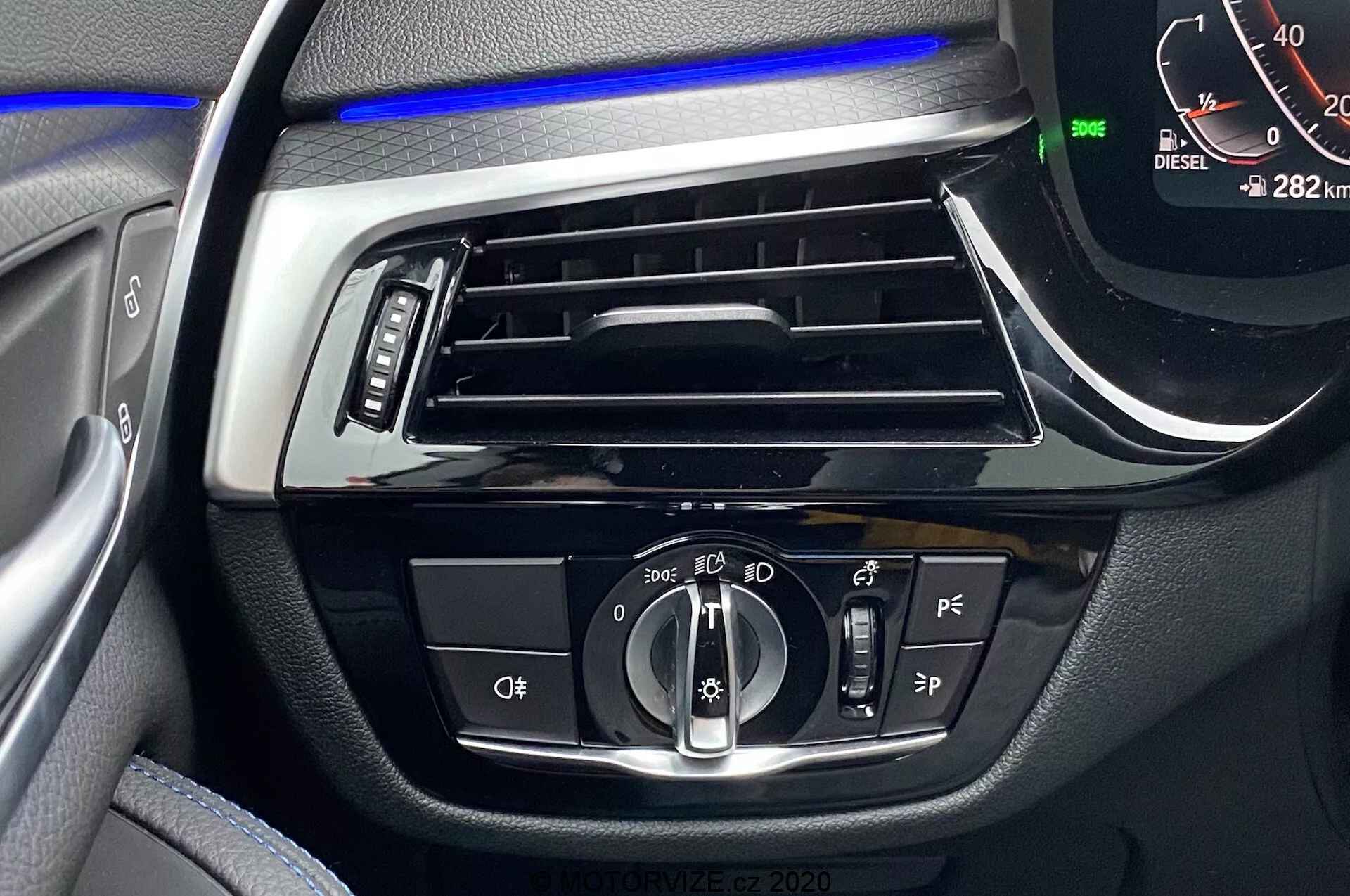 Gros plan sur l'intérieur de la BMW Série 5 2020, se concentrant sur le tableau de bord côté conducteur avec une bande de lumière ambiante bleue au-dessus de la ventilation horizontale. Les commandes centrales incluent un bouton rotatif pour les réglages automatiques et des phares, des boutons-poussoirs probablement pour les feux antibrouillard et le nivellement des phares, des boutons d'assistance au conducteur supplémentaires et un compteur de vitesse numérique affichant 0 km/h avec le véhicule en mode Parking et une lecture de 282 km. Un indicateur 'DIESEL' est également visible.