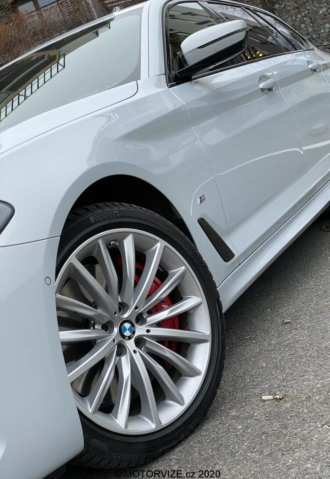 Vue latérale en gros plan d'une BMW 520i Touring Steptronic (2020) blanche, centrée sur la roue avant en alliage moderne à multiples rayons avec le logo BMW visible et l'étrier de frein rouge, montrant partiellement le profil latéral avec une ligne de caractère nette à travers les poignées des portes, un rétroviseur latéral de la même couleur que la voiture avec signal de virage intégré et un petit badge sur le panneau latéral avant. L'éclairage naturel assure une visibilité claire des caractéristiques du véhicule. Photo de MOTORVIZE.cz 2020.
