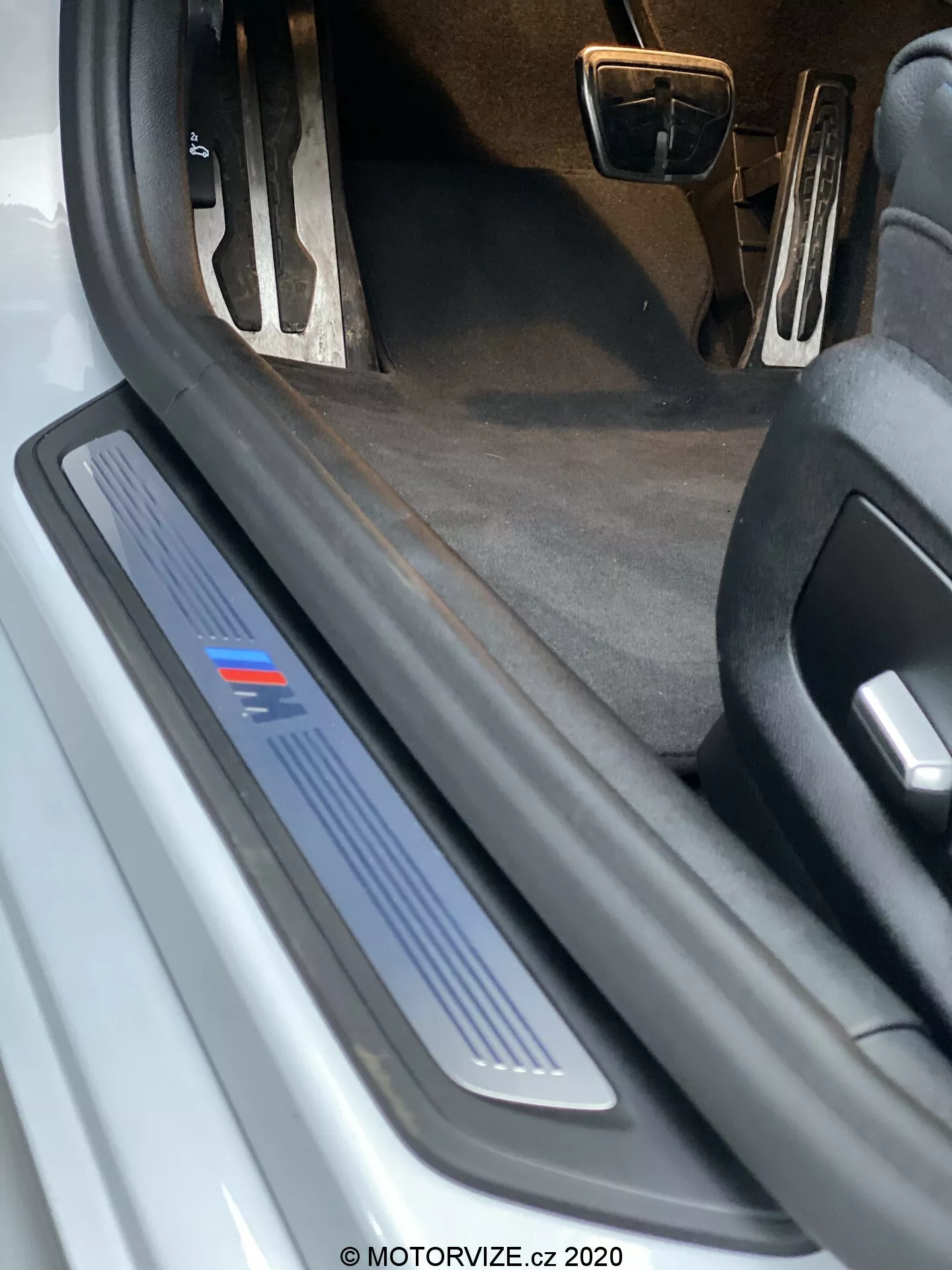 Vue en gros plan du seuil de porte côté conducteur d'une BMW 520i Touring Steptronic 2020. L'image montre une perspective en contre-plongée, mettant en avant la plaque de seuil en métal brossé avec une bande bleue, qui pourrait indiquer un niveau de finition spécifique ou une édition sportive. La porte ouverte n'est pas visible, mais la plaque de seuil protectrice et décorative est évidente, suggérant une possible illumination LED. Le tapis intérieur sombre et la vue partielle des pédales avec finition métallique indiquent un possible pack intérieur sportif. Il y a des débris et des signes d'usure visibles sur la plaque de seuil, ce qui suggère que la voiture est utilisée.