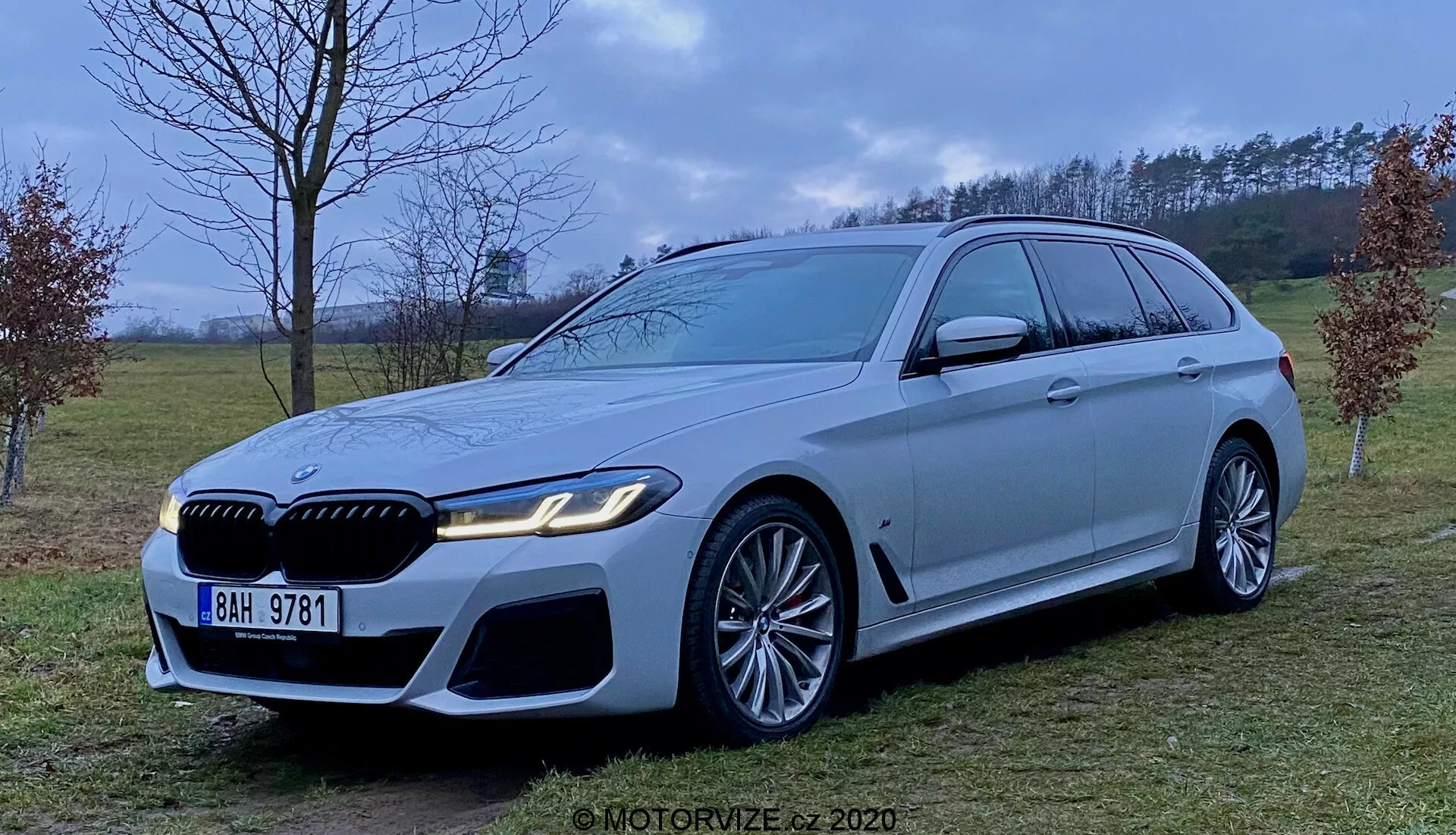 Image d'un break de luxe BMW Série 5 Touring 2020 (BMW 520i Touring Steptronic) de couleur argentée ou grise, présentant une face avant agressive et sportive avec de grandes prises d'air, une calandre proéminente et des phares LED élégants. Le véhicule est équipé de grandes jantes en alliage à plusieurs rayons et d'étriers de frein remarquables, une longue ligne de toit avec un spoiler, des garnitures chromées autour des fenêtres et une ligne d'épaule marquée. Photographie prise à mi-hauteur dans un environnement semi-rural avec une lumière diffuse.
