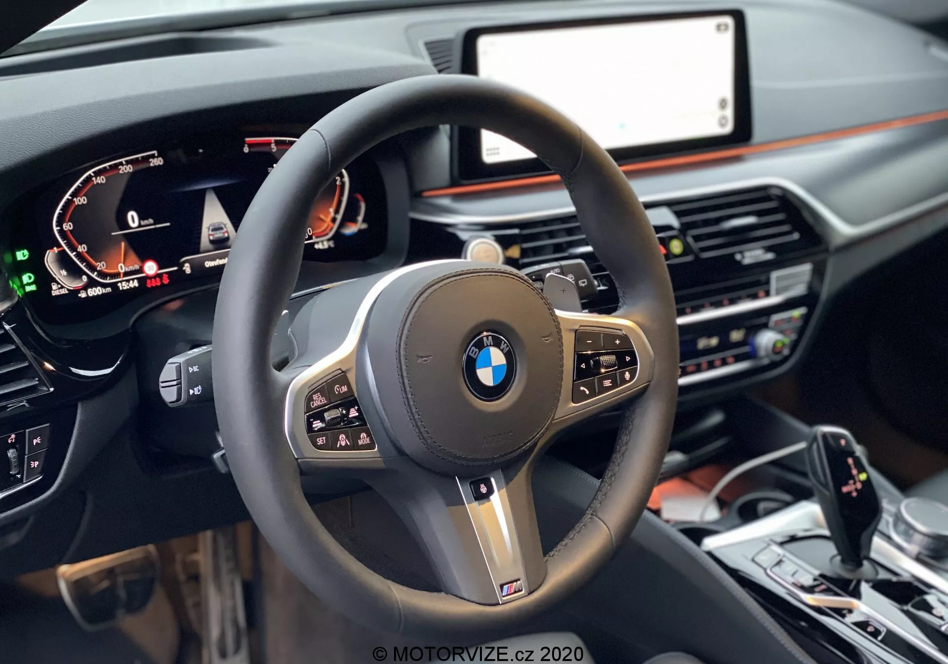 Vue à la première personne depuis le siège conducteur d'une BMW 520i Touring Steptronic 2020, centrée sur le volant moderne à trois branches avec une jante épaisse en cuir, des boutons multifonctions et un logo BMW proéminent au centre, entouré de finitions noires brillantes et d'accents métalliques. Un tableau de bord numérique à gauche affiche la vitesse, le régime moteur et les informations du conducteur, tandis qu'à droite, partiellement visible, se trouve l'écran central d'infodivertissement. Le tableau de bord aux tons sombres avec une touche de bois intègre les commandes de climat et le système multimédia sous l'écran. En bas de l'image, on voit la console centrale avec le sélecteur de vitesses, le contrôleur iDrive et les boutons de contrôle de la dynamique de conduite.