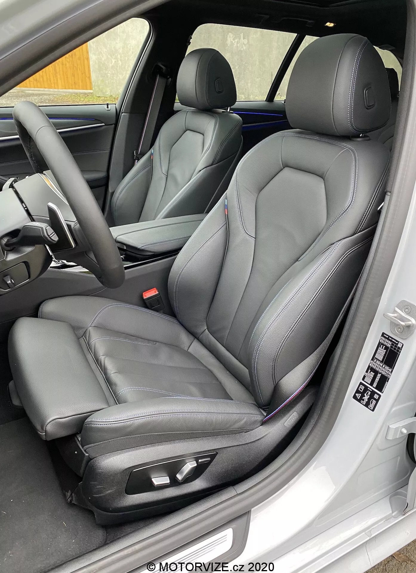 Vue rapprochée et de côté du siège conducteur de la BMW 520i Touring Steptronic 2020, montrant des sièges avant modernes de type baquet en cuir noir avec surpiqûres contrastées, avec des commandes de réglage électroniques pour un confort accru. L'image capture également le panneau intérieur de la porte conducteur avec commandes des vitres et des rétroviseurs électriques, réglages de la mémoire du siège et autres caractéristiques du tableau de bord et du volant indiquant luxe et technologie avancée.