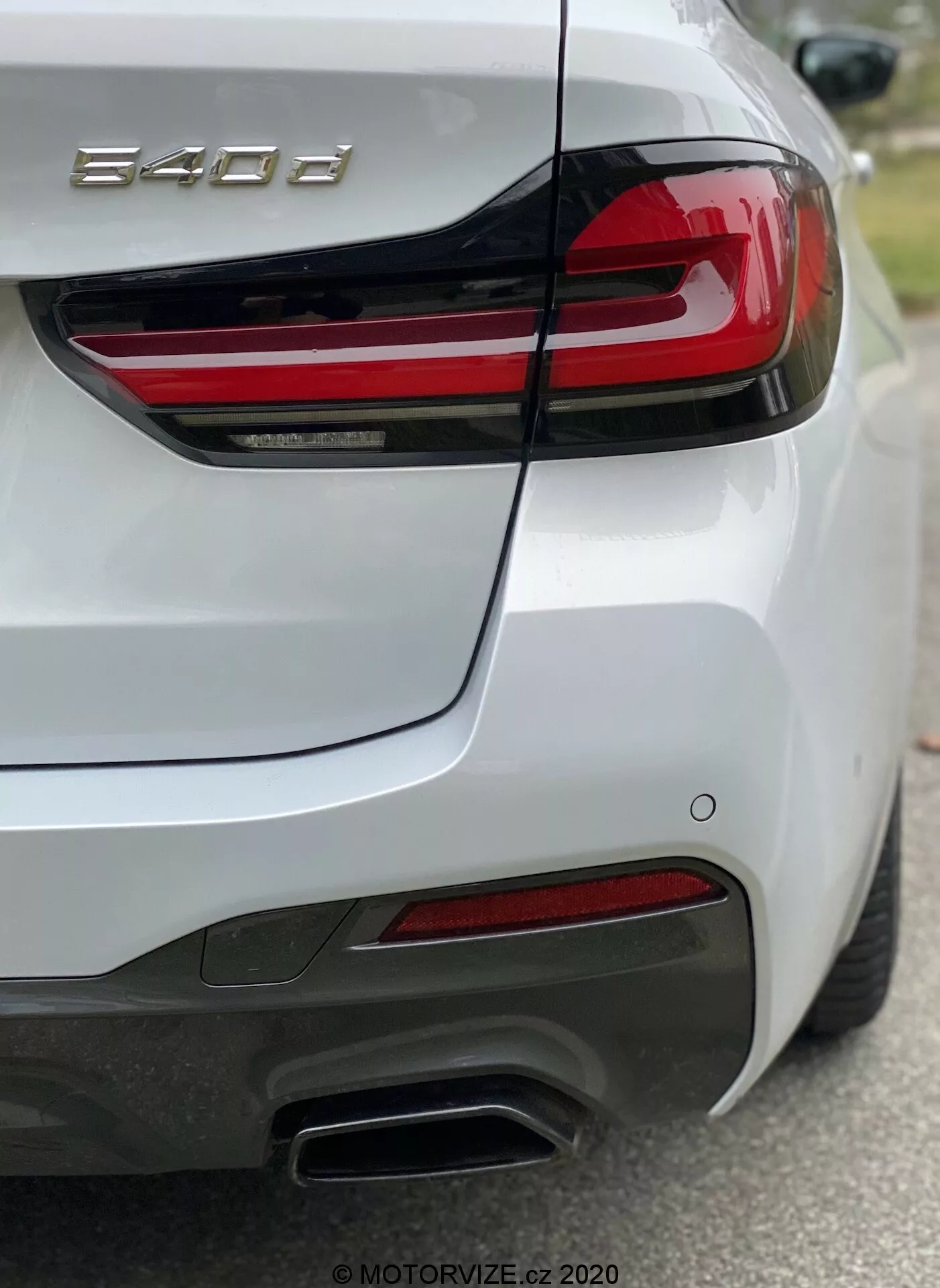 Vue arrière d'une BMW Série 8 (840i), capturée de l'angle arrière-droit, mettant en valeur le feu arrière droit avec des bandes LED rouges horizontales, le couvercle du coffre sculpté avec spoiler intégré, le pare-chocs arrière avec capteurs, les doubles embouts d'échappement métalliques du côté droit, les réflecteurs rouges et la peinture blanche brillante. La photo se concentre sur le badge '840i', montrant les détails de design et les caractéristiques technologiques telles que l'éclairage et les systèmes d'assistance au conducteur.