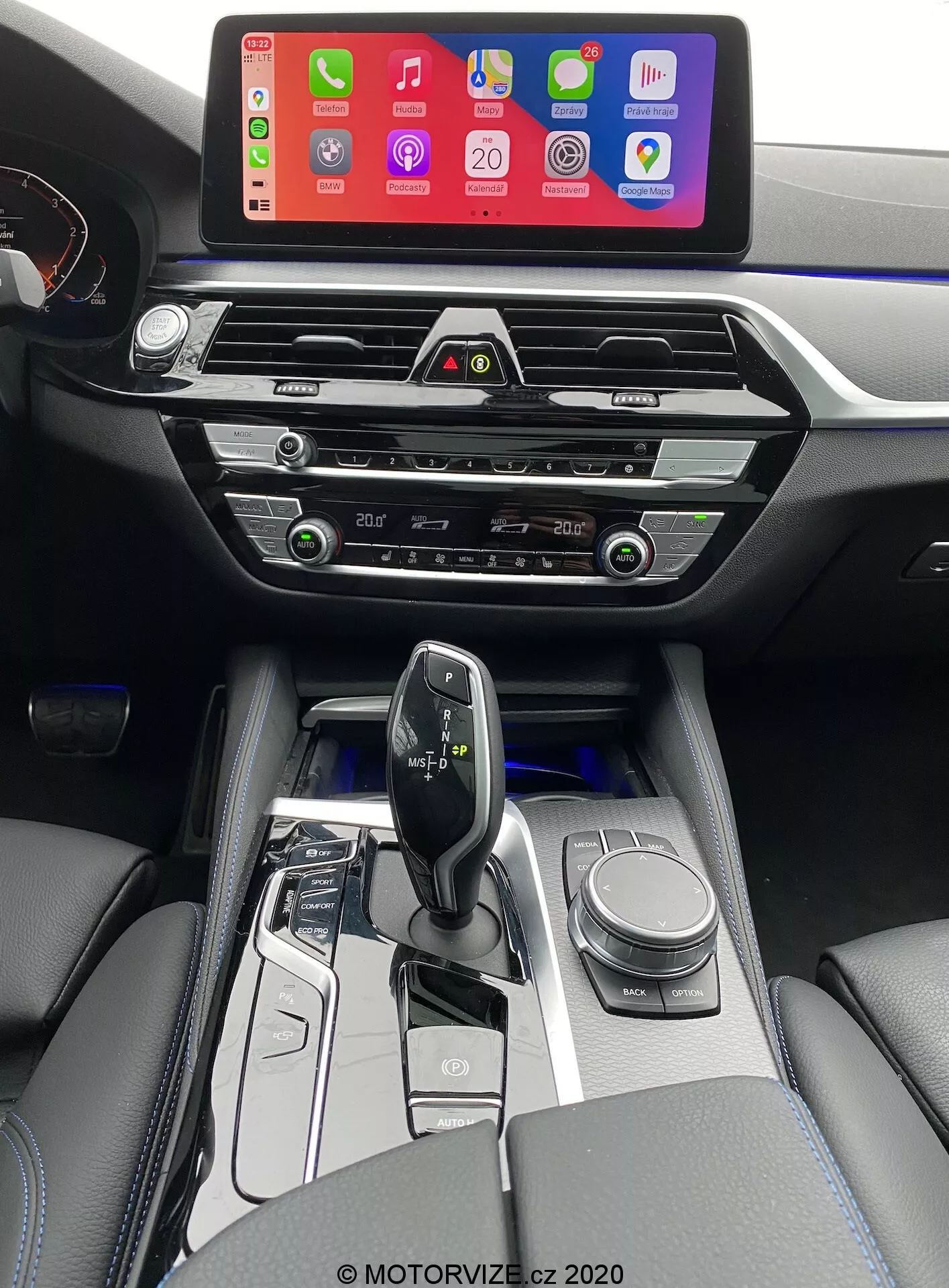 Photo intérieure d'une BMW 520i Touring 2020 avec transmission Steptronic, prise de la perspective du conducteur et légèrement surélevée pour offrir une vue plus claire de la console centrale, de la zone du changement de vitesse et d'une partie du tableau de bord. L'image montre un grand écran d'infodivertissement affichant une interface de connexion pour smartphone, probablement Apple CarPlay, monté en hauteur sur le tableau de bord pour une visibilité facile. Sous l'écran se trouvent des bouches d'aération horizontales modernes avec des accents chromés, suivies de boutons et de cadrans tactiles pour le contrôle du climat. La console centrale abrite également les commandes du système audio, des commandes climatiques supplémentaires et une éventuelle fente pour lecteur CD. La manette de changement de vitesse ergonomique et minimaliste, probablement électronique, est entourée de garnitures en noir brillant et de accents métalliques, avec des boutons de sélection du mode de conduite et un bouton rotatif de navigation. L'intérieur comprend également des accoudoirs en cuir avec des coutures visibles, ajoutant une touche de luxe. Un éclairage blanc doux met en évidence les boutons autour de la zone du levier de vitesses, indiquant une transmission automatique.