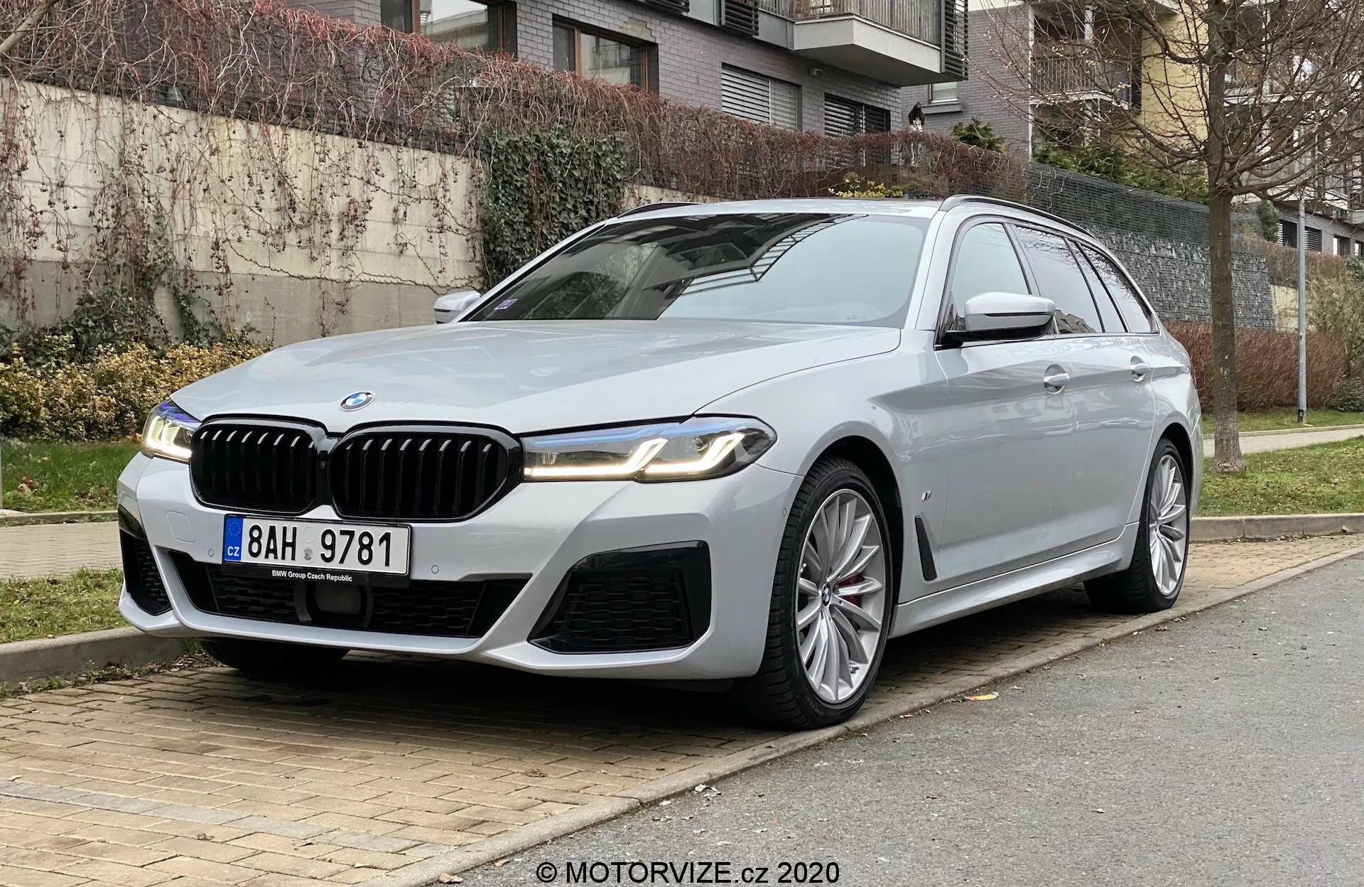 BMW 520i Touring de luxe garée dans une rue de la ville, vue sous un angle frontal trois-quarts bas mettant en valeur l'avant et le côté conducteur. La voiture se distingue par une grille de calandre proéminente avec des détails chromés, des phares LED élégants, un pare-chocs avant sportif et des plis définis sur le capot. Les jantes en alliage à plusieurs branches, les rétroviseurs latéraux avec clignotants et caméra, les cadres de fenêtres chromés et un toit ouvrant ajoutent à l'apparence luxueuse. Positionnée sur le côté droit avec de l'herbe, des arbustes et des bâtiments résidentiels en arrière-plan, suggérant une technologie avancée et un design haut de gamme.