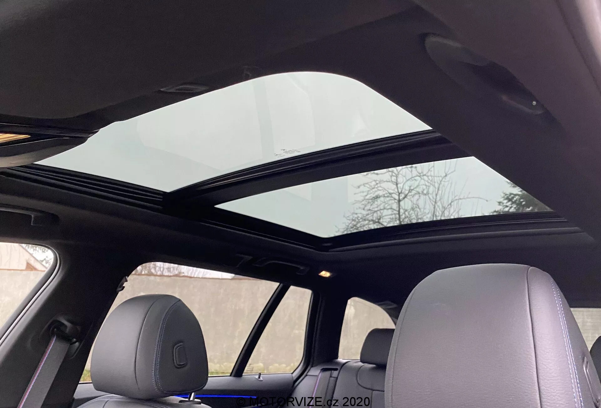 Vue intérieure d'une BMW Série 5 2020 520i Touring avec transmission Steptronic, montrant un toit ouvrant panoramique s'étendant sur les sièges avant et arrière du point de vue d'un passager à l'arrière regardant vers le haut. Le panneau avant du toit ouvrant est légèrement ouvert, améliorant la ventilation. Le ciel de toit est de couleur claire, complétant le design intérieur cohérent, avec des appuie-têtes assortis au matériau intérieur, soulignant le confort avancé et les commodités modernes.