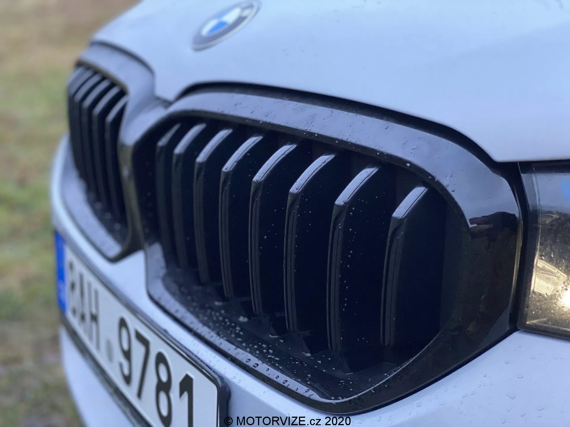 Gros plan de la calandre avant d'une BMW 520i Touring Steptronic de l'année 2020, montrant la calandre distinctive en forme de rein et l'emblème BMW avec des lamelles verticales noires, un phare partiel et des gouttelettes d'eau suggérant une pluie récente ou un lavage, prise sous un angle bas depuis l'avant-gauche.