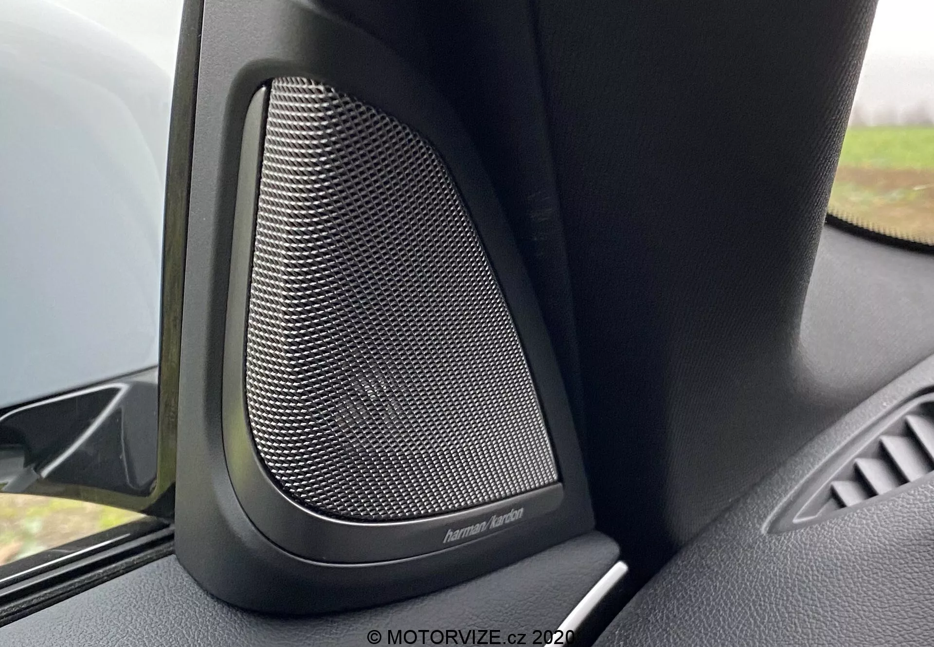 Vue en gros plan de l'intérieur de la BMW 5er-Reihe 2020 (BMW 520i Touring Steptronic) se concentrant sur le haut-parleur de porte premium Harman Kardon. L'image capture le motif en maille fine de la grille métallique du haut-parleur, entourée de matériaux en plastique noir ou de type soft-touch conçus pour amortir les vibrations et offrir une qualité sonore supérieure. L'angle de la caméra montre la partie supérieure du panneau de porte, avec le haut-parleur en premier plan et une petite partie de l'accoudoir de porte. Un flou doux de l'arrière-plan met en évidence cette caractéristique audio haut de gamme dans ce véhicule de luxe.