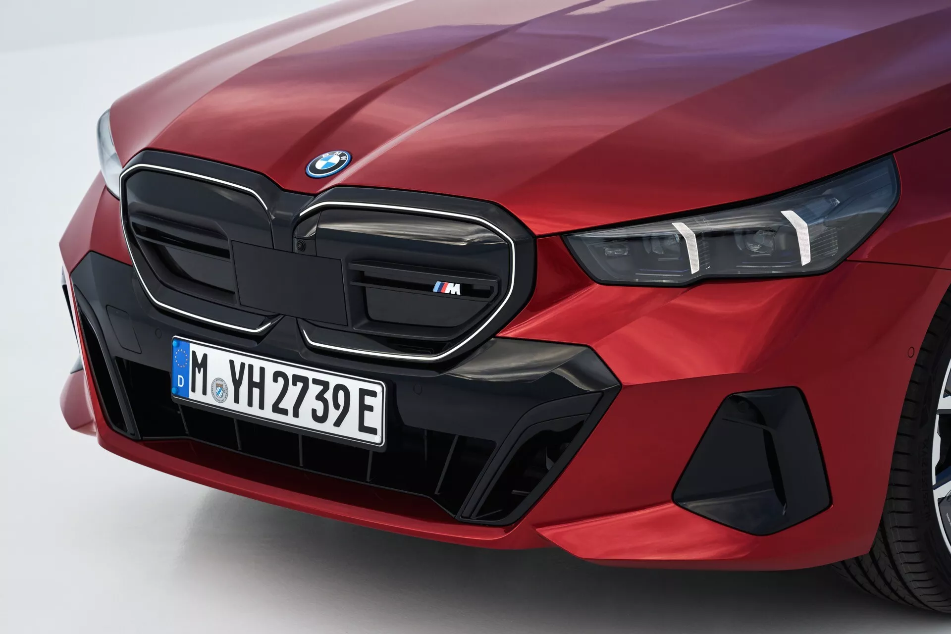 Vue rapprochée et angulaire du côté avant gauche d'une BMW Série 5 (G60-G61) 2023 en rouge, présentant la grille rénale emblématique marquée comme un modèle 'M', un bloc phare LED et des éléments aérodynamiques spécialisés suggérant des capacités électriques ou hybrides