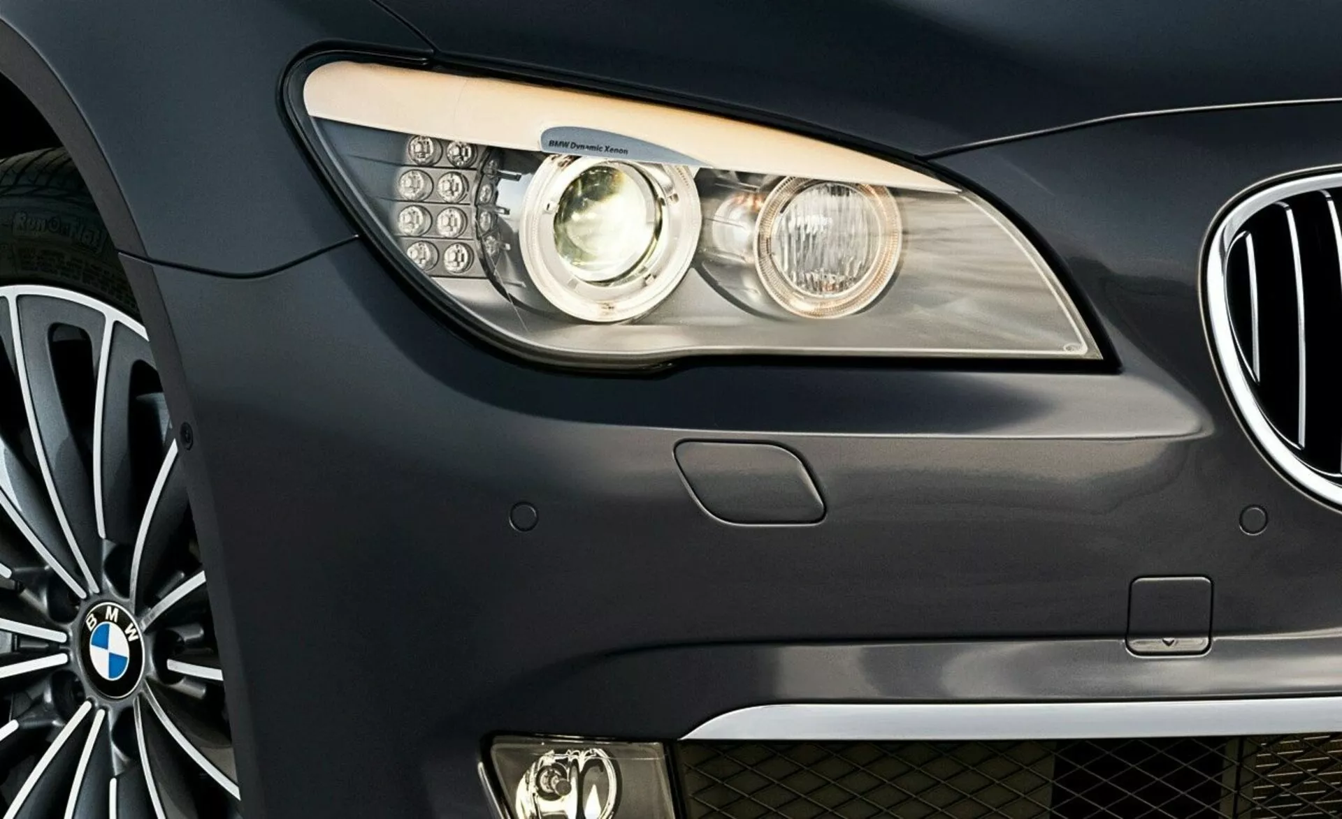 Vue latérale avant d'une BMW Série 7 de 2005 avec l’emblème de la marque, l’ensemble de phares modernes avec lentille de projecteur et feux de jour LED, vue partielle de la calandre en forme de rein et roue en alliage à plusieurs branches.