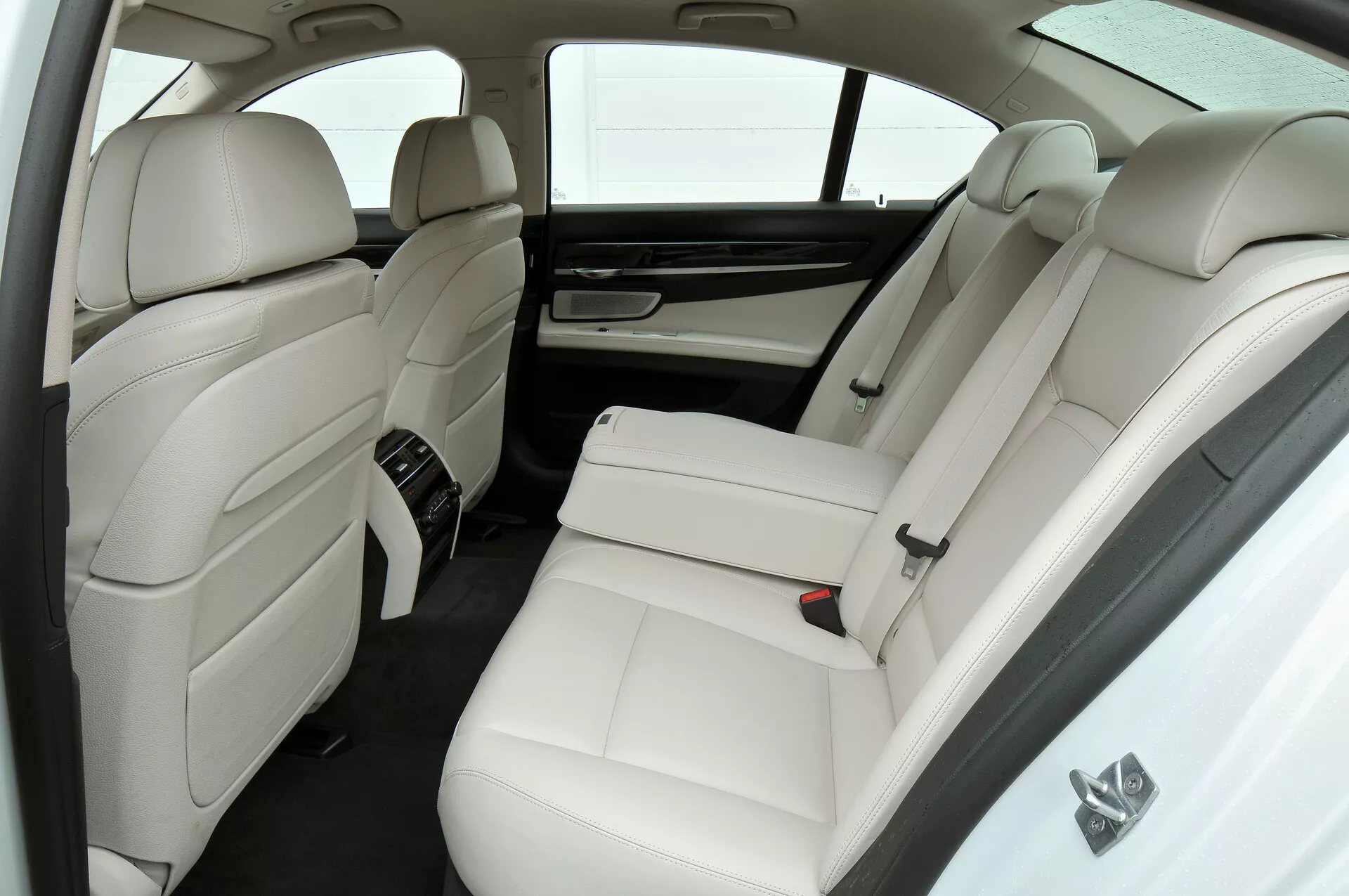 L'image présente l'intérieur luxueux en cuir blanc d'une BMW Série 7 (facelift 2012, F01-F02) en se concentrant sur le compartiment arrière des passagers depuis une perspective légèrement élevée. Elle comprend deux sièges arrière séparés avec appuie-têtes et renforts latéraux, une console centrale avec commandes, des bouches d'aération de climatisation arrière, un pare-soleil sur la fenêtre arrière, des appuie-têtes intégrés aux dossiers des sièges avant et une palette de couleurs minimaliste en blanc et tons foncés.