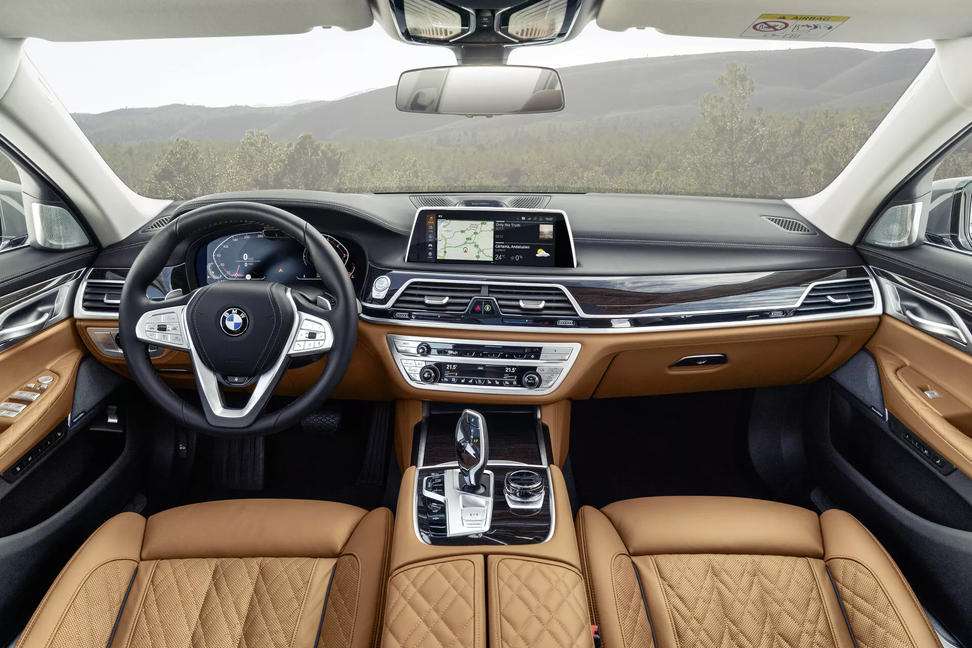 L'image montre l'intérieur d'une BMW Série 7 de 2019 (facelift G11/G12) depuis la perspective des sièges arrière, mettant en avant le design détaillé et luxueux de l'habitacle avant. Dotée d'un volant moderne avec des boutons multifonctions et un emblème BMW distinct, le tableau de bord élégant comporte un écran central tactile pour l'infodivertissement. La console centrale comprend un levier de vitesses automatique, un pavé tactile et divers systèmes de contrôle dynamique, complétés par des sièges en cuir beige matelassé indiquant un niveau de finition premium. Les panneaux des portes reflètent ce luxe avec du cuir beige, des garnitures en bois et un éclairage d'ambiance, suggérant que des fonctionnalités technologiques avancées et de confort sont intégrées dans tout le véhicule, y compris un toit ouvrant pour plus de lumière naturelle.