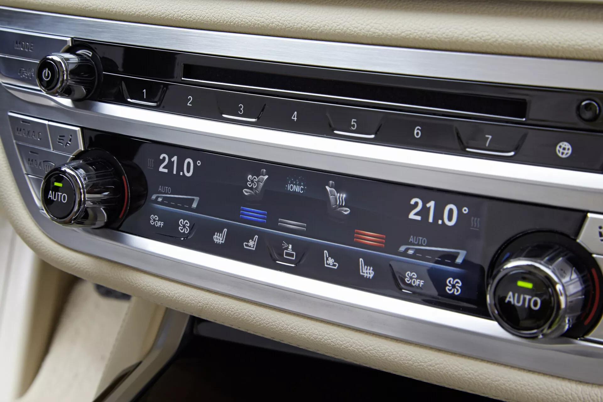 Vue rapprochée du système de contrôle climatique de la BMW 7er-Reihe 2015 (G11-G12) sur la console centrale entre les sièges du conducteur et du passager. Se concentre sur le panneau horizontal avec deux boutons et des boutons pour diverses fonctions. Les deux boutons sont réglés à 21,0°C et marqués 'AUTO', suggérant un contrôle automatique de la température à deux zones. Les boutons comprennent le dégivrage de la lunette arrière, la circulation de l'air, le dégivrage du pare-brise et l'activation des évents. Le système est encadré de garnitures métalliques brossées contre un intérieur de couleur claire, avec une bande noire, peut-être un écran ou une surface tactile, juste au-dessus.