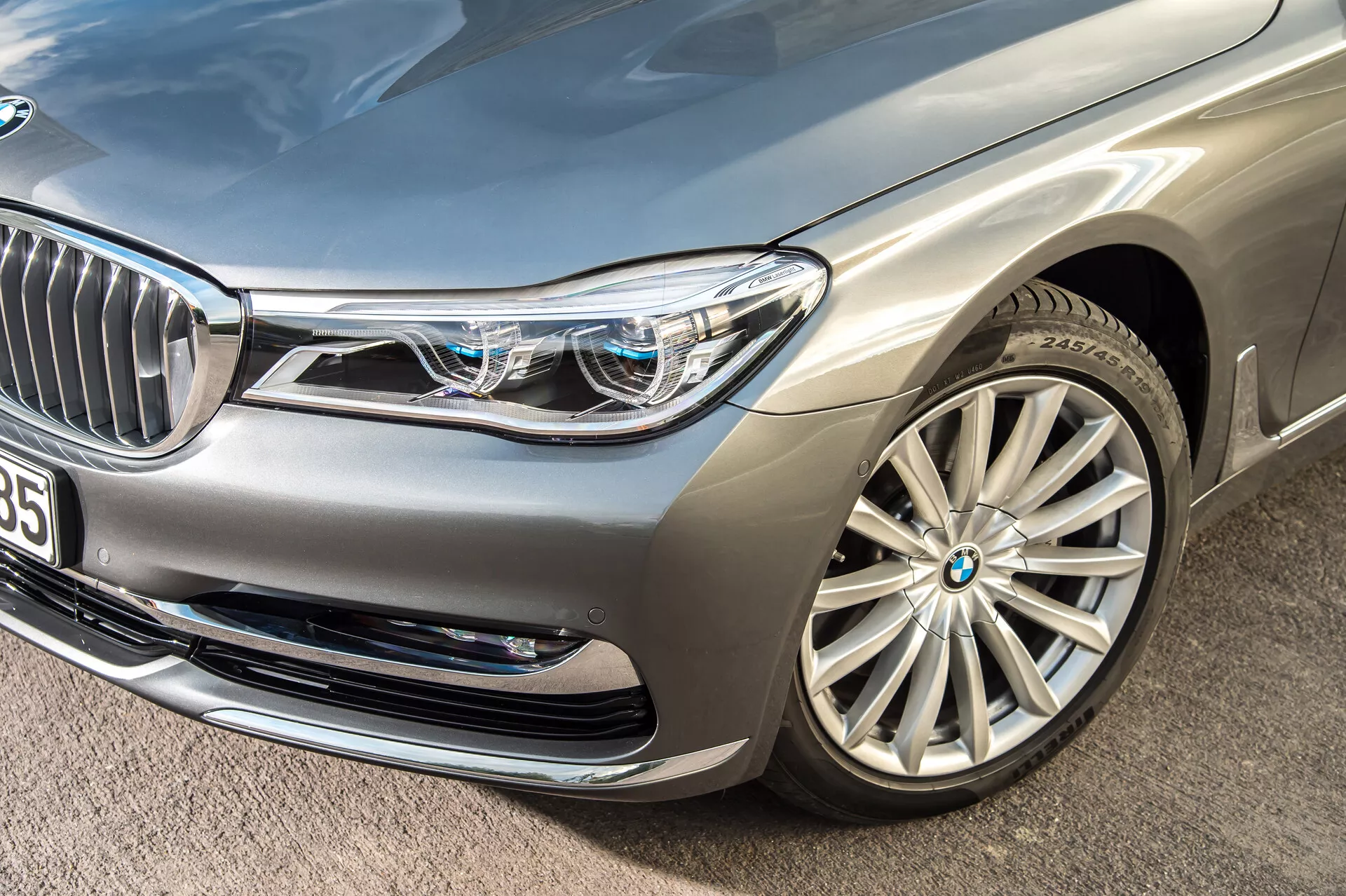 Photo en gros plan en biais d'une berline de luxe argentée BMW 7er-reihe 2015 (G11-G12), centrée sur la section avant gauche. Points saillants: le pare-chocs avant, le phare gauche, le capot, l'aile et la roue en alliage multi-branches sont visibles. Phares LED complexes avec feux diurnes distinctifs et petits carrés bleus indiquant des systèmes d'éclairage avancés. La calandre avant partiellement visible avec des lamelles verticales chromées et une caméra ou un capteur à proximité suggèrent une technologie d'assistance à la conduite. Les prises d'air sur le pare-chocs avant et la finition de peinture métallique mettent encore plus en valeur le design luxueux et aérodynamique de la voiture.