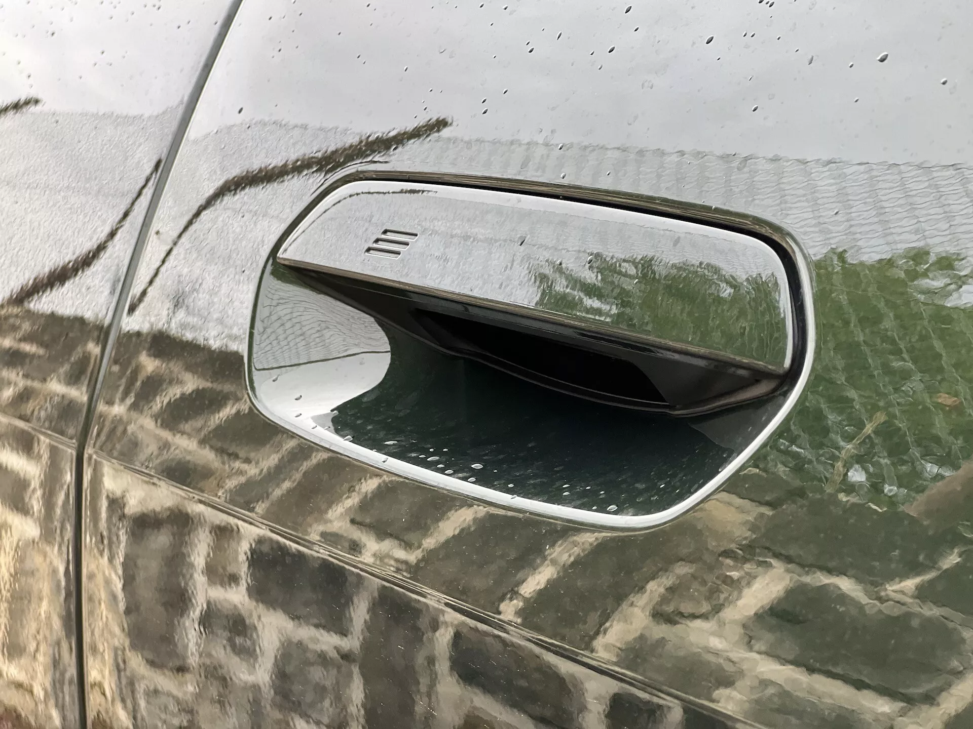 Vue rapprochée du rétroviseur côté conducteur de la BMW i4 2021 montrant son design aérodynamique, son boîtier noir avec une texture fine en ligne horizontale, sa bordure argentée, sa surface résistante aux intempéries avec des gouttes d'eau et sa fonctionnalité pliable reflétée dans le verre moucheté de pluie.