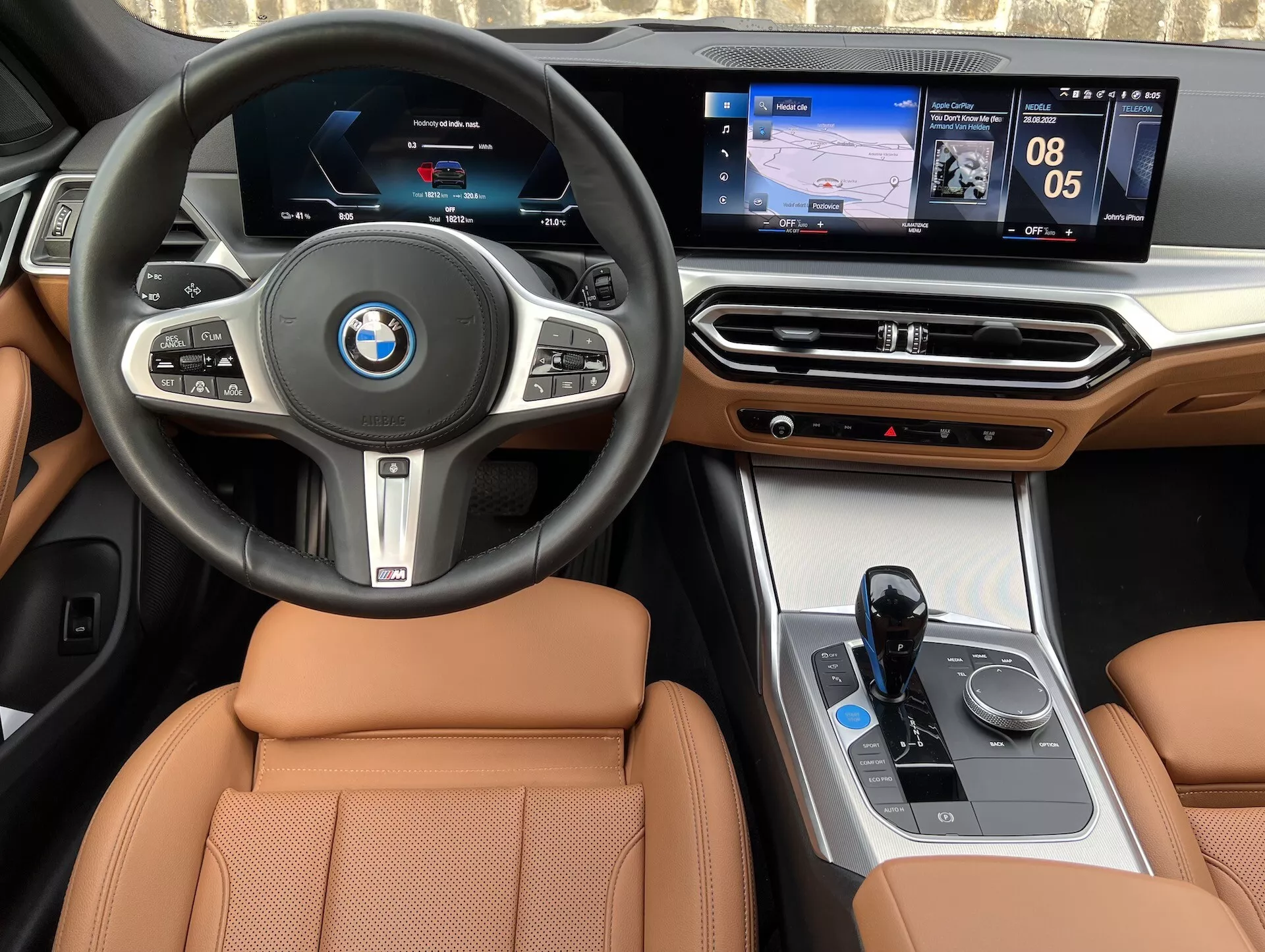 Vue intérieure d'une BMW i4 2021, montrant un volant central avec logo BMW, boutons multifonctions, cluster d'instruments numériques, système d'infodivertissement central tactile affichant navigation et médias, accents de bois de luxe sur le tableau de bord, interface de contrôle climatique, sélecteur de vitesse électronique, contrôleur iDrive et sièges en cuir couleur tan.