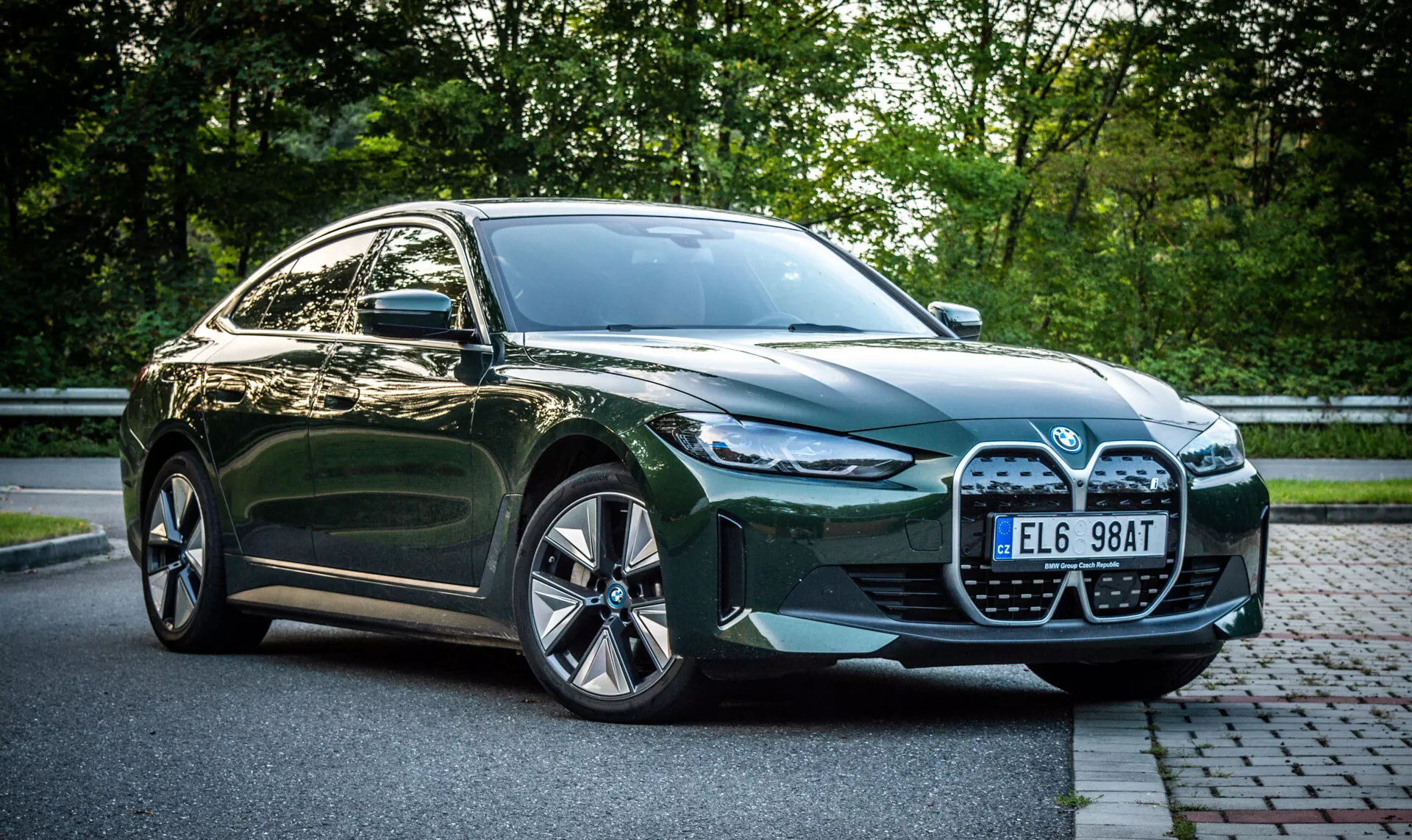 Berline BMW i4 verte quatre portes de 2021 garée sur une rue pavée à côté d'une zone gazonnée avec du feuillage en arrière-plan, vue de quart avant mettant en avant la grande calandre, les phares LED, les prises d'air et les jantes en alliage