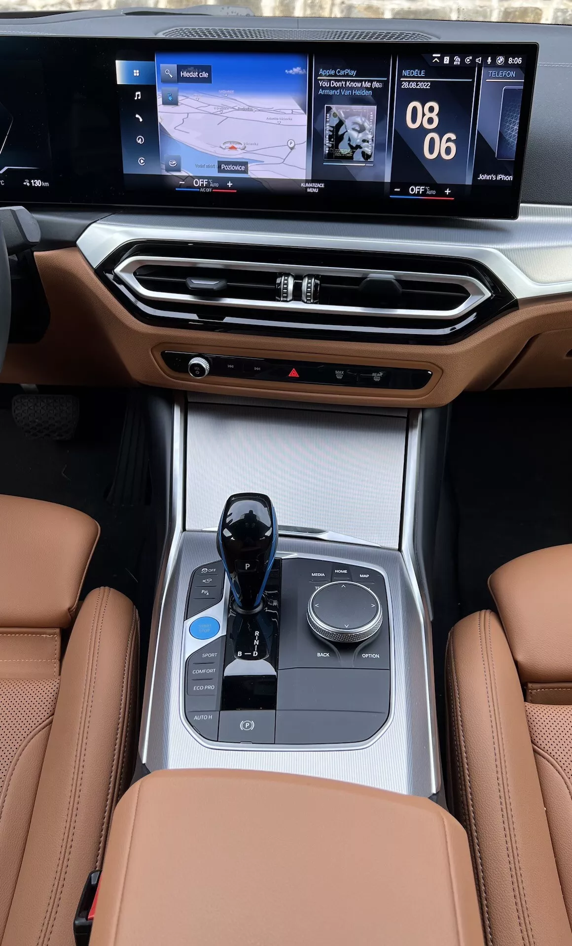 Vue de la console centrale intérieure d'une BMW i4 2021 depuis les sièges arrière, mettant en évidence les sièges en cuir, le sélecteur de vitesse électronique, le bouton de contrôle multimédia, les porte-gobelets, les commandes climatiques et l'écran d'infodivertissement à double interface avec navigation et affichage média