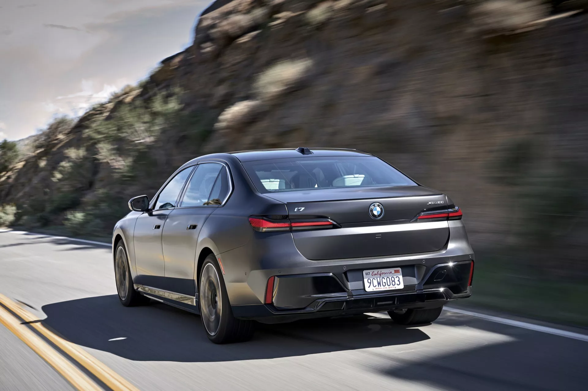 Sedan moderne BMW i7 (G70) de 2022 en mouvement sur une autoroute, présentant sa silhouette sportive de type coupé, des feux arrière LED distinctifs à travers le hayon, de grandes roues en alliage et un système d'échappement, en fini sombre brillant avec des vitres teintées