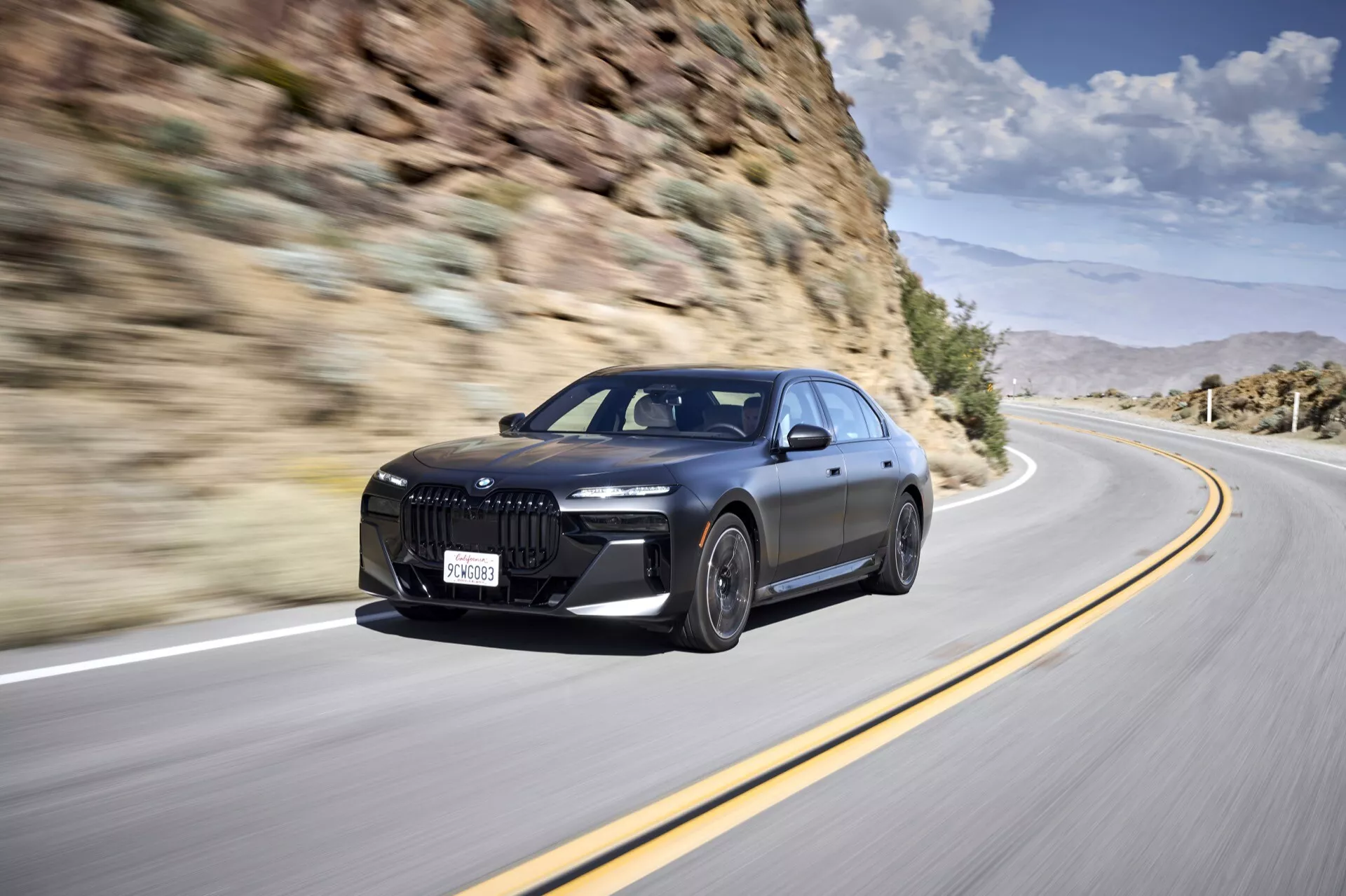 Berline de luxe BMW i7 (G70) 2022 en mouvement, mettant en avant sa grande grille avant distinctive, phares LED, pare-chocs avant aérodynamique avec de grandes entrées d'air, lignes sportives sur le capot, grandes roues en alliage et profil latéral élégant avec poignées de porte affleurantes, sur fond de paysage montagneux.