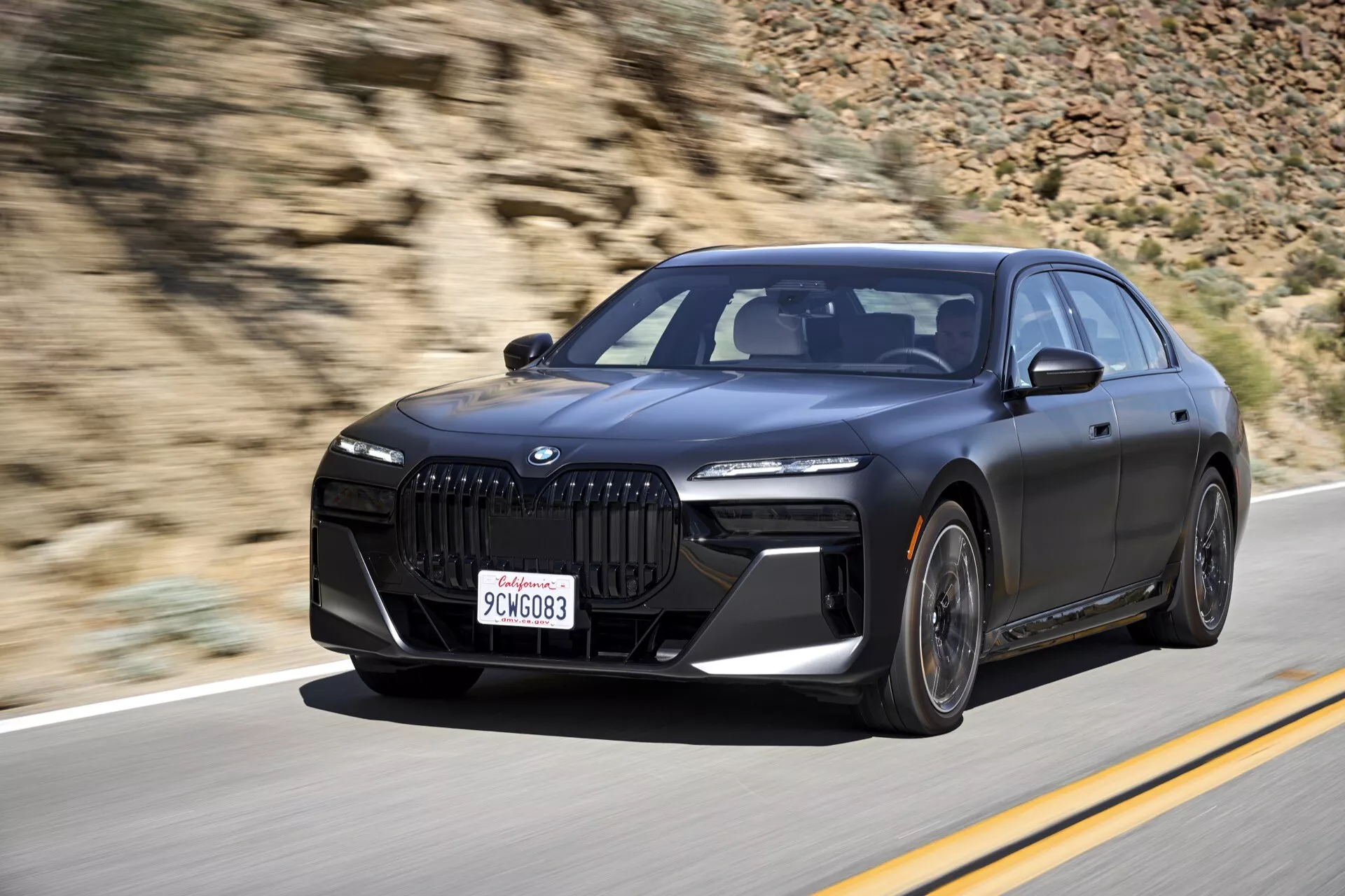 Sedan de luxe BMW i7 G70 2022 en mouvement, présentant son design élégant et ses caractéristiques modernes telles que la calandre en forme de rein et les phares LED d'une vue de trois quarts avant sur un fond de mouvement flou