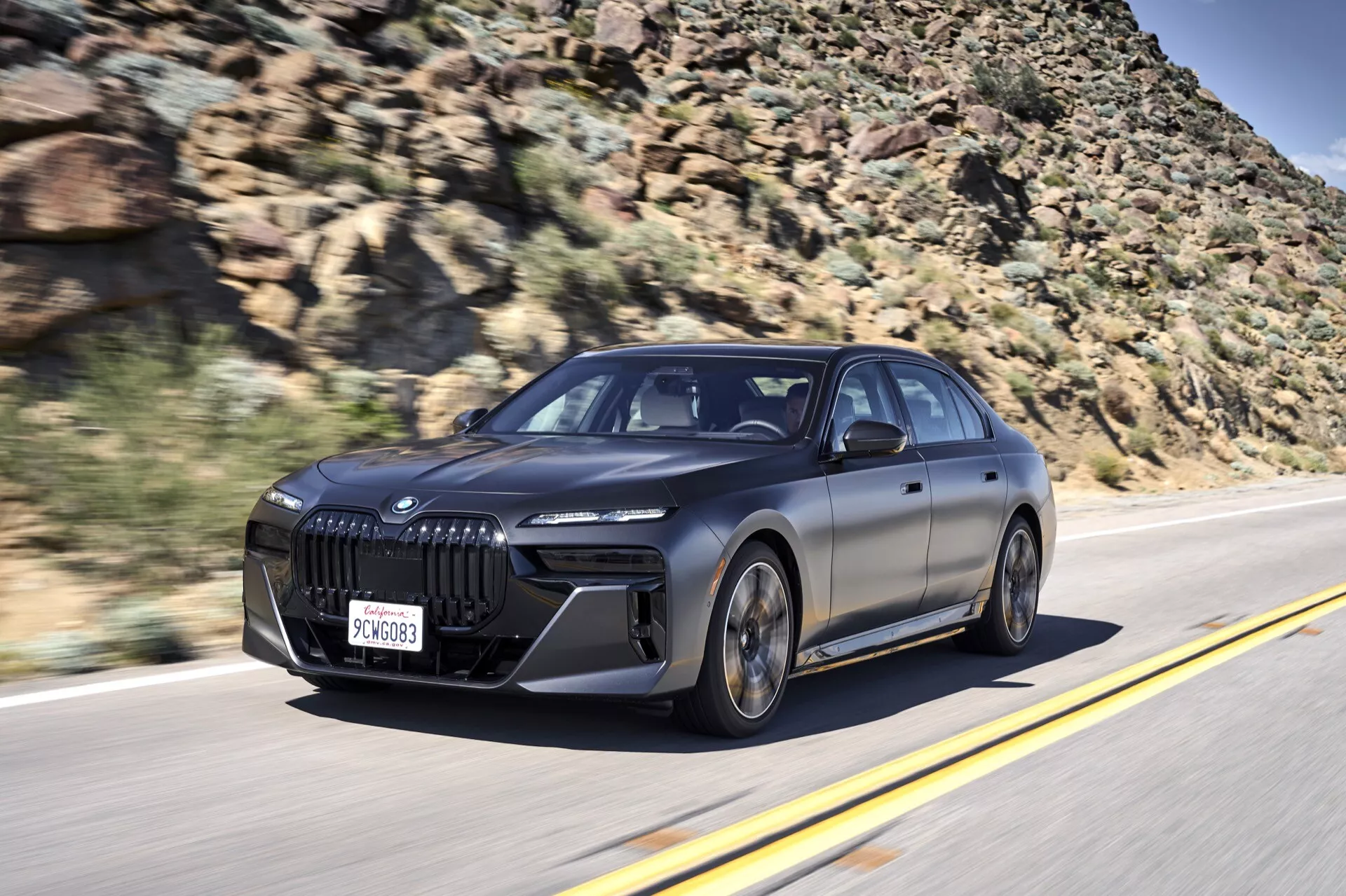 Vue dynamique avant trois quarts de la BMW i7 (G70) 2022 en mouvement, soulignant sa grande grille en forme de rein, ses phares LED et ses caractéristiques de design élégantes sur un arrière-plan de route floue et de paysage rocheux.