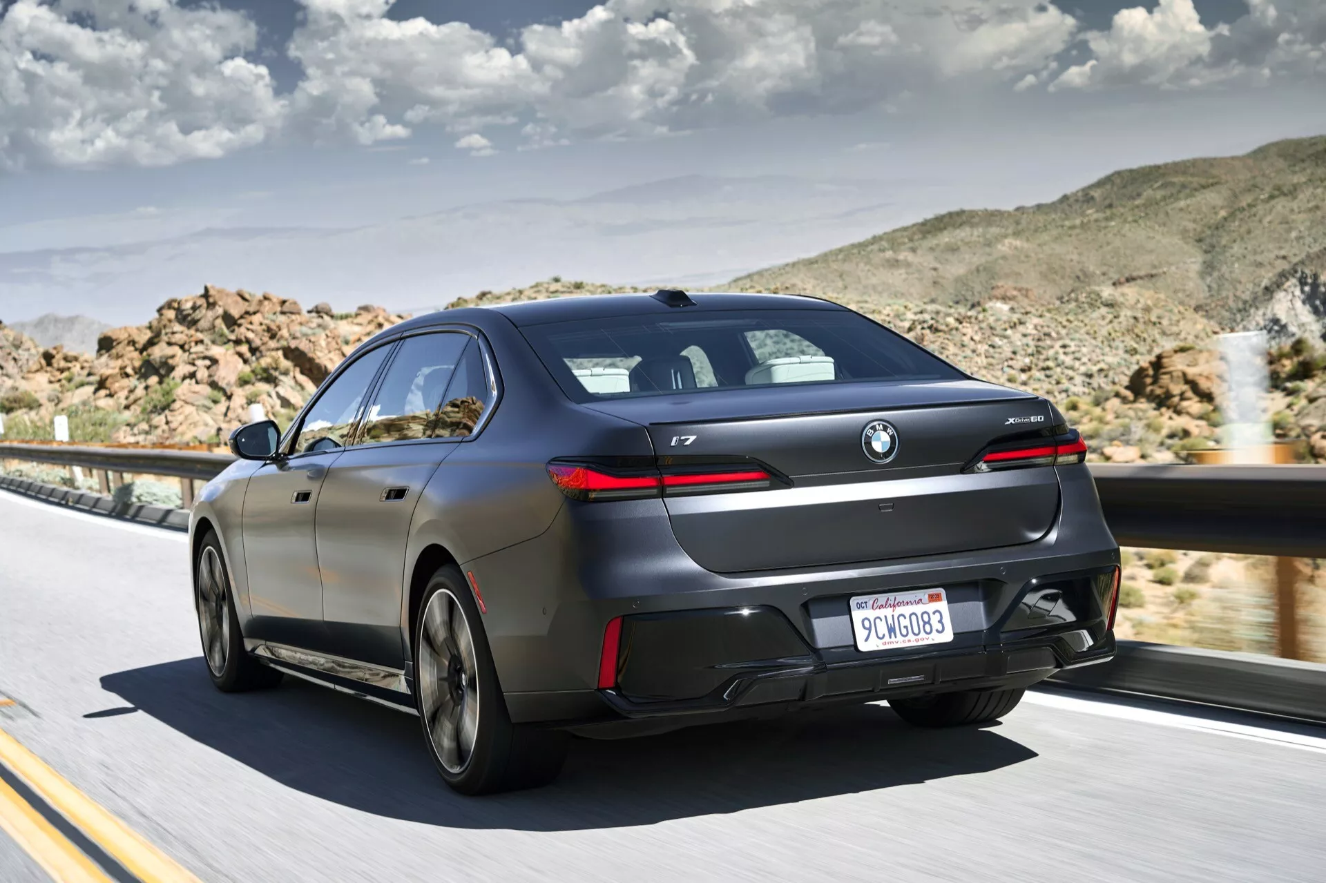 BMW i7 (G70) 2022 en mouvement, vue arrière trois quarts mettant en évidence les feux arrière, le pare-chocs arrière et les lignes aérodynamiques latérales contre un paysage désertique