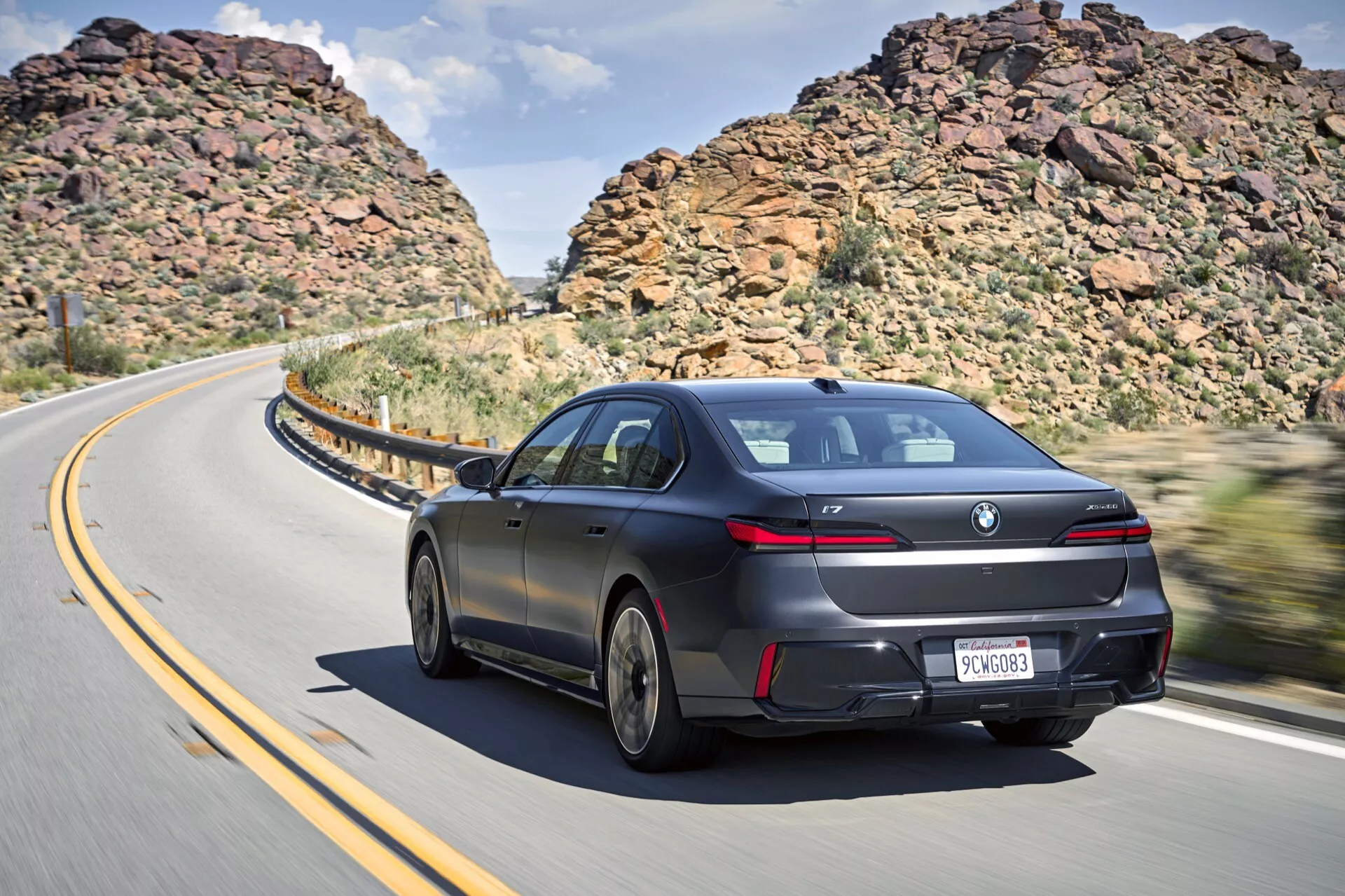 Vue arrière dynamique trois quarts d'une berline BMW i7 2022 (G70) en mouvement sur une route courbe avec des collines escarpées et un ciel partiellement nuageux, présentant un design aérodynamique élégant, une peinture lustrée, des jantes en alliage de designer, des feux arrière LED, un spoiler arrière subtil, des vitres teintées et des sorties d'échappement doubles.