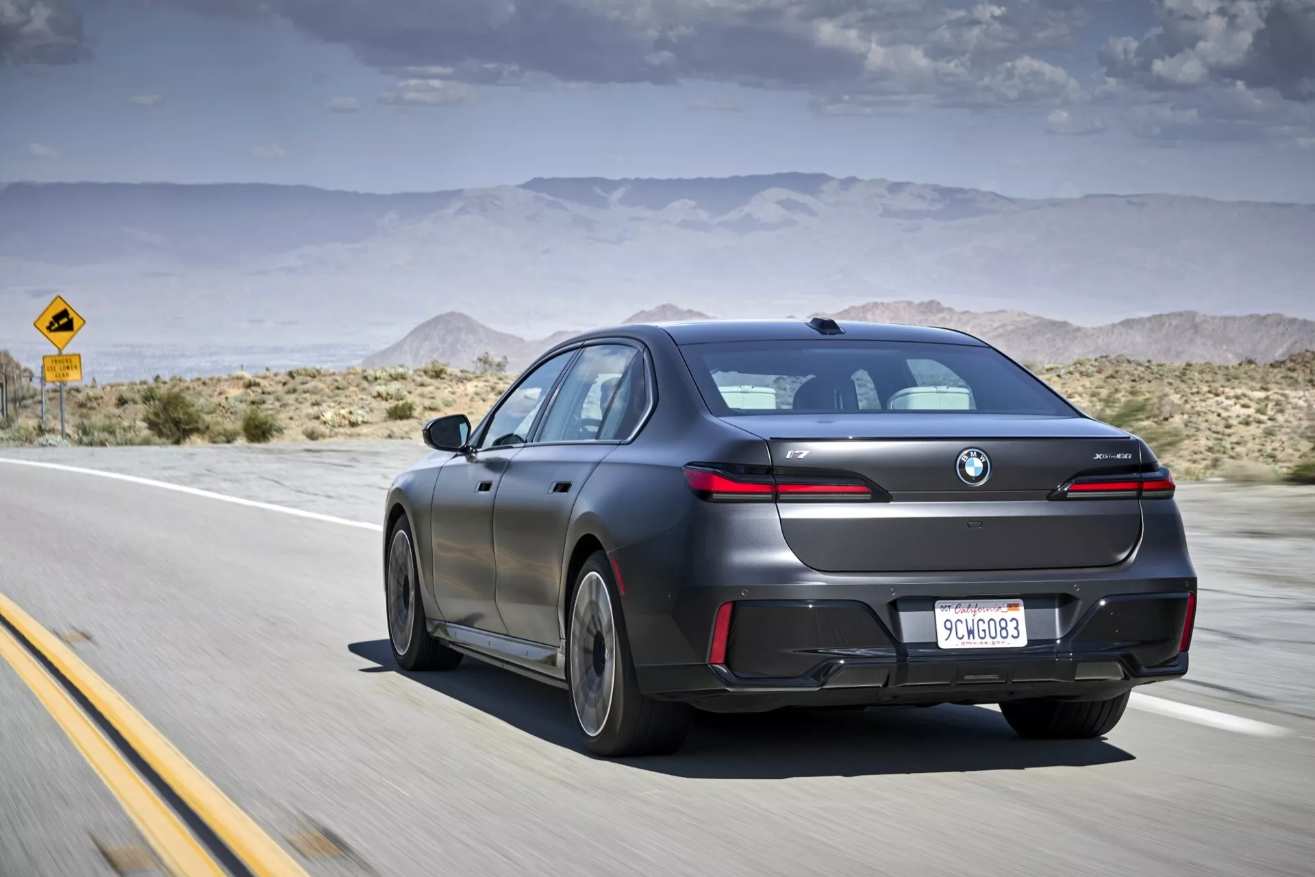 Séduisante berline moderne BMW i7 (G70) de 2022 en finition mate, capturée en mouvement depuis un angle extérieur arrière de trois quarts sur une autoroute désertique, mettant en avant son design aérodynamique, ses feux arrière à LED et ses caractéristiques technologiques telles que des capteurs de stationnement et un toit ouvrant, sous un ciel partiellement nuageux.