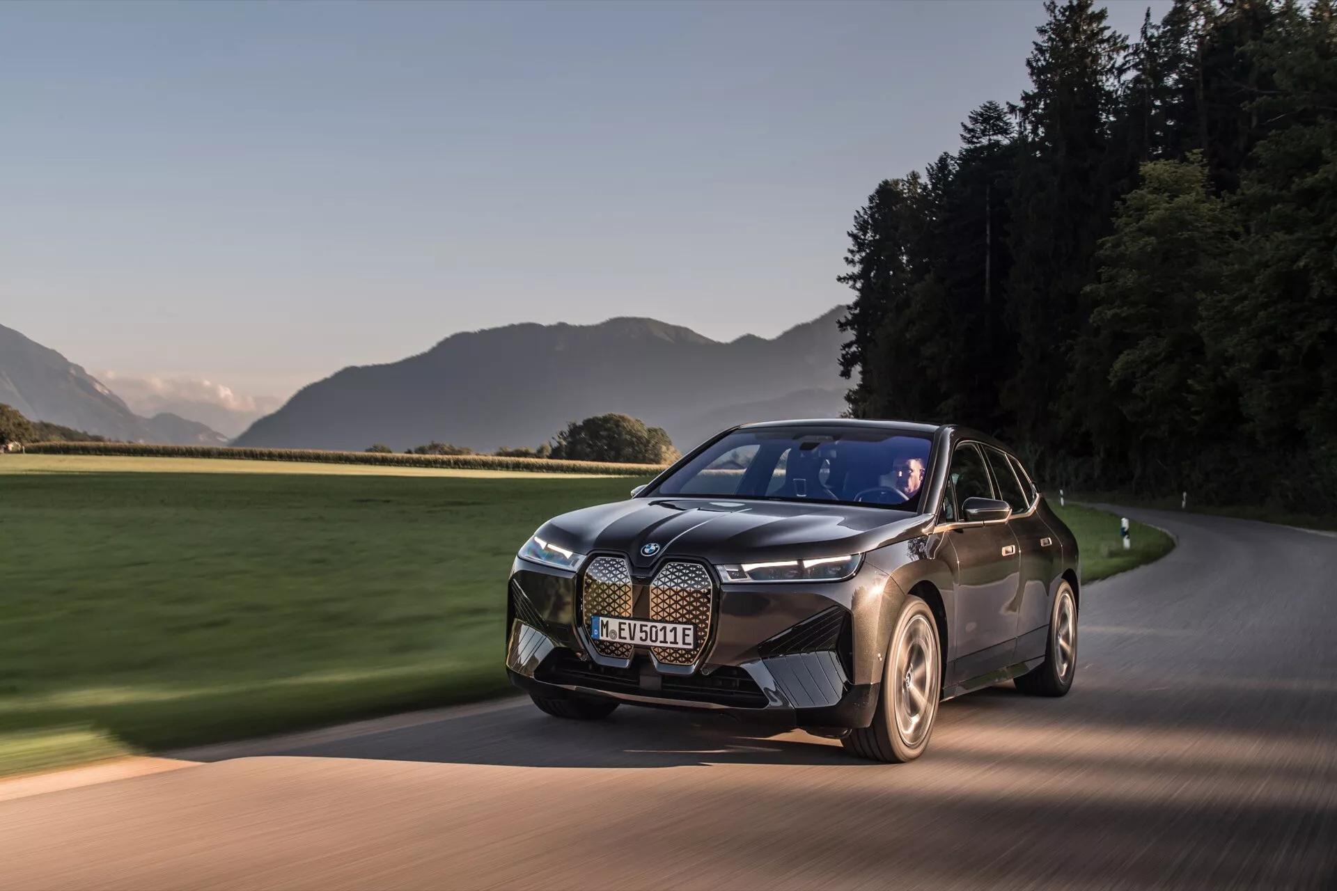 SUV électrique moderne BMW iX 2021 (i20) en mouvement sur une route pavée à l'aube ou au crépuscule, présentant sa peinture bicolore, son design élégant avec une grille proéminente, ses phares distinctifs et ses roues aérodynamiques sur fond de paysage pittoresque.