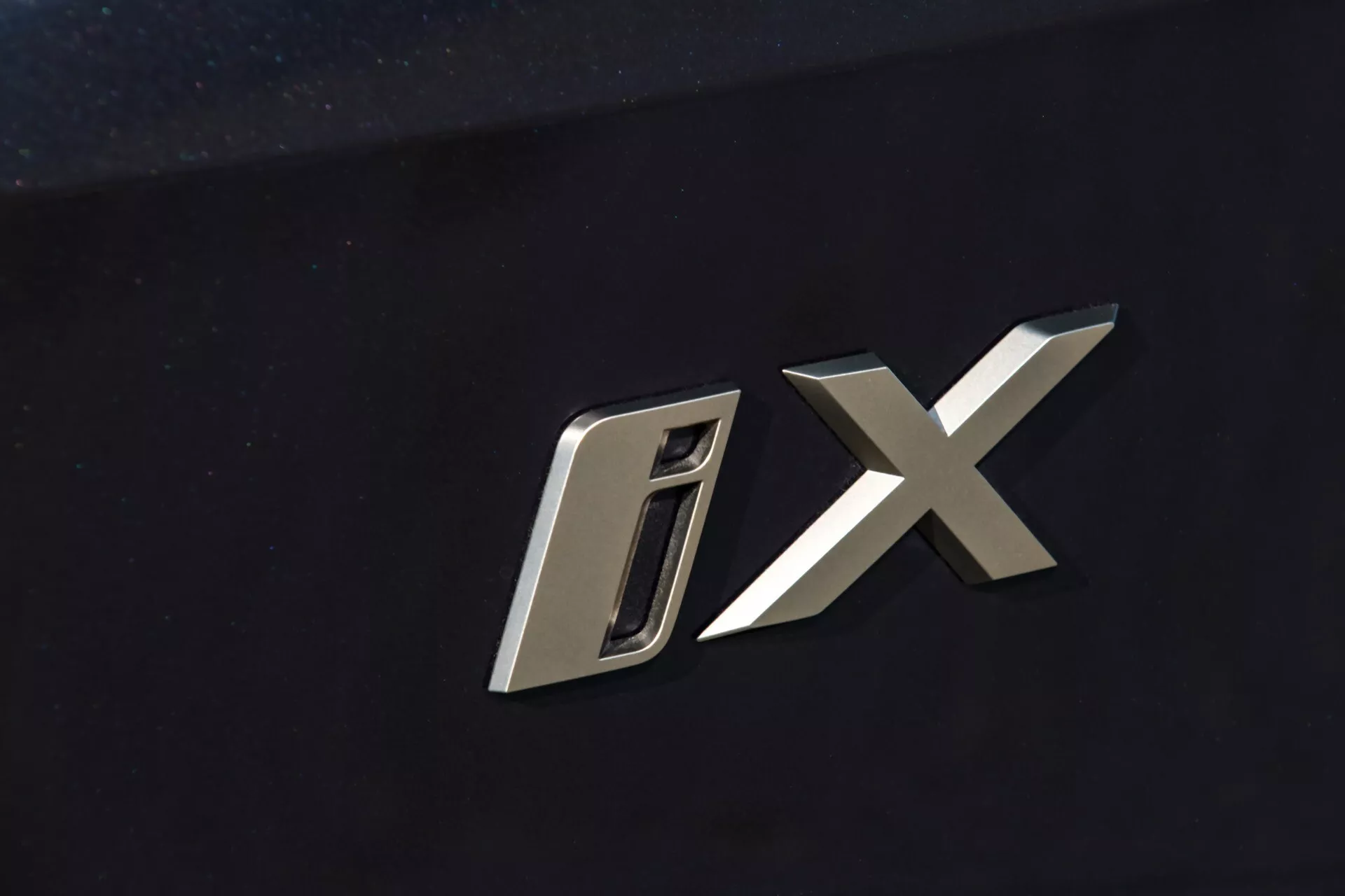 Vue rapprochée de l'emblème du modèle BMW iX 2021 avec caractères métalliques stylisés 'D' et 'X' sur un fond sombre de carrosserie