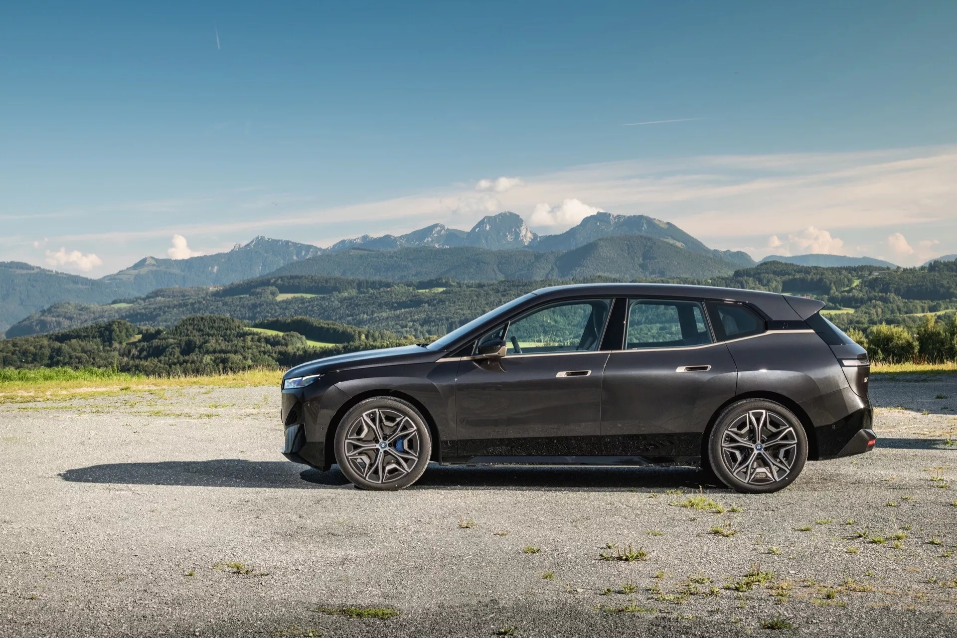 Profil latéral d'un SUV moderne BMW iX (i20) 2021 sur gravier avec arrière-plan montagneux sous un ciel bleu clair, doté de grandes roues en alliage à rayons multiples, d'un design élégant avec une ligne de ceinture haute et un toit incliné, de vitres teintées, de clignotants intégrés dans les rétroviseurs de couleur carrosserie, d'une grille avant visible et d'un bloc phares LED, de garnitures chromées et d'un toit panoramique.