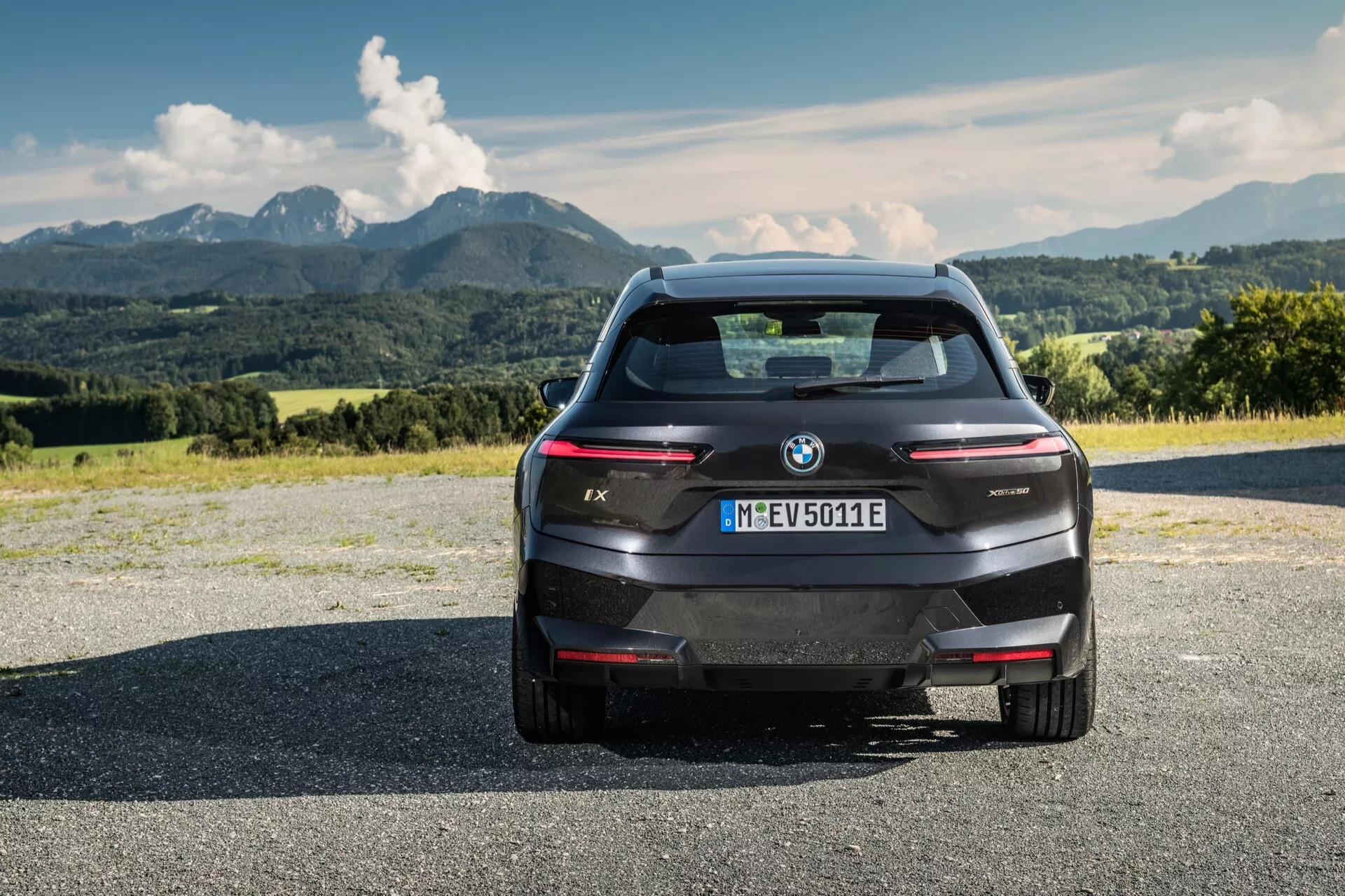 Vue arrière d'un SUV électrique BMW iX 2021 sur une surface en asphalte avec des montagnes et un ciel partiellement nuageux en arrière-plan, mettant en avant son design moderne, ses lignes élégantes, le logo BMW et la désignation du modèle, les capteurs de stationnement arrière intégrés, le feu stop surélevé, la caméra de recul, les feux arrière horizontaux et fins et une fenêtre arrière aérodynamique sans échappement traditionnel, indiquant sa nature électrique et son immatriculation à Munich.