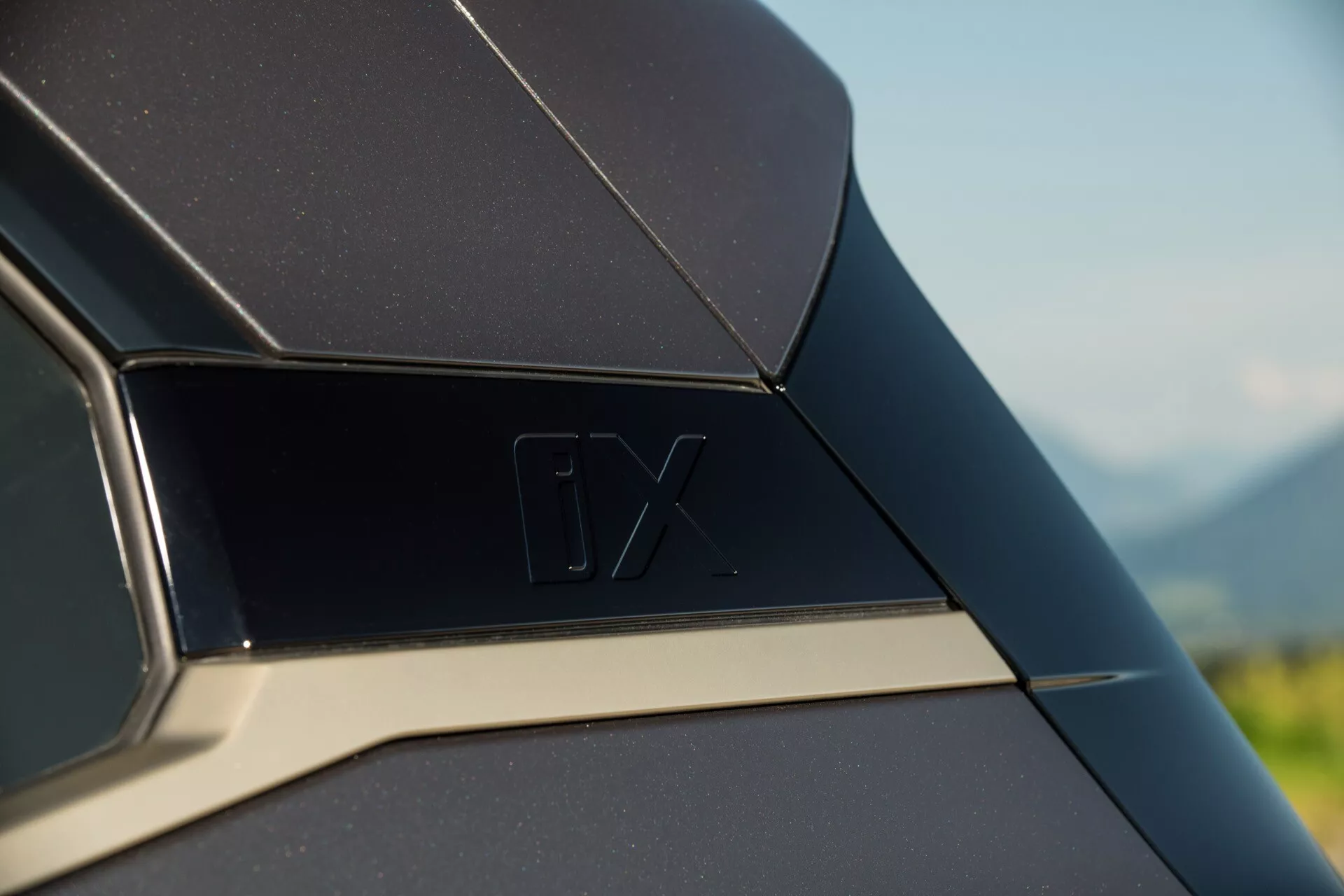 Image en gros plan d'un détail extérieur du BMW iX 2021, mettant en vedette le panneau arrière ou la zone du montant C avec l'insigne 'iX' et une ligne de garniture chromée contrastante, sur un fond naturel flou