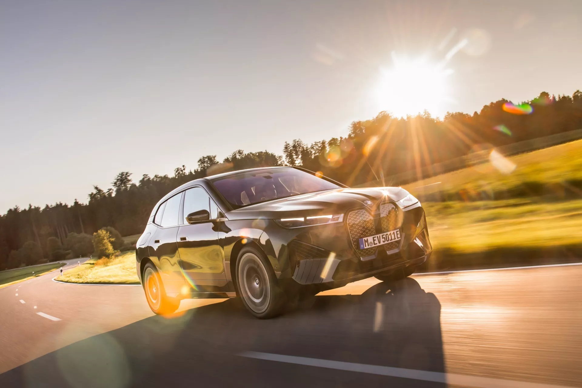 Vue avant dynamique aux trois quarts d'une BMW iX 2021 en mouvement, mettant en évidence sa stance sportive, ses phares LED élégants et sa grille distinctive et grande, prise depuis un angle bas pour souligner les caractéristiques de design moderne et agressif de la voiture contre un arrière-plan naturel rural.