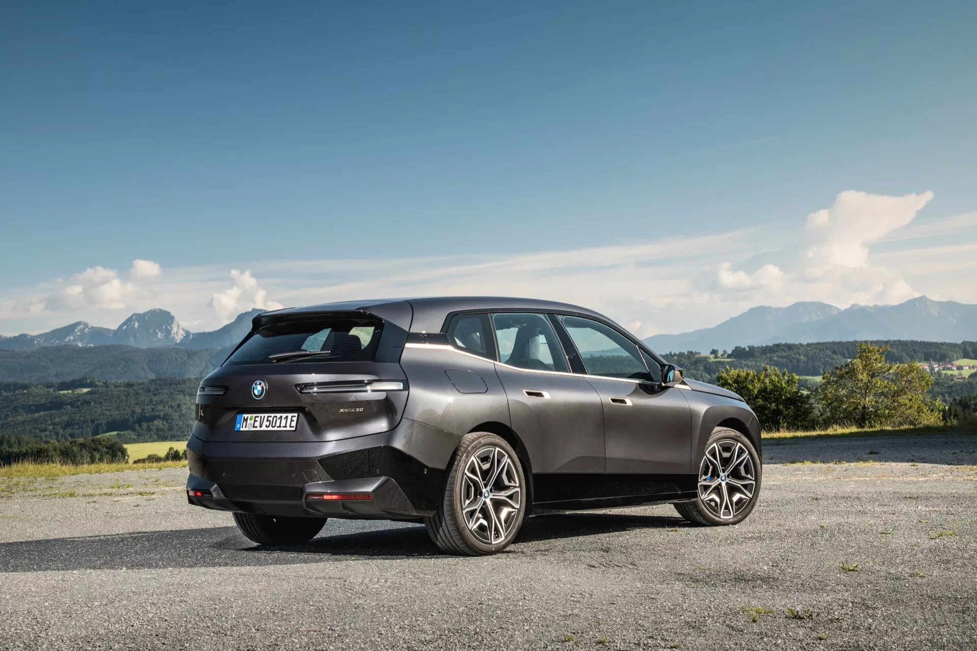 Vue arrière trois quarts d'un BMW iX 2021 en gris foncé, garé sur une surface pavée surélevée avec un paysage montagneux en arrière-plan, reflétant un design moderne de SUV de luxe