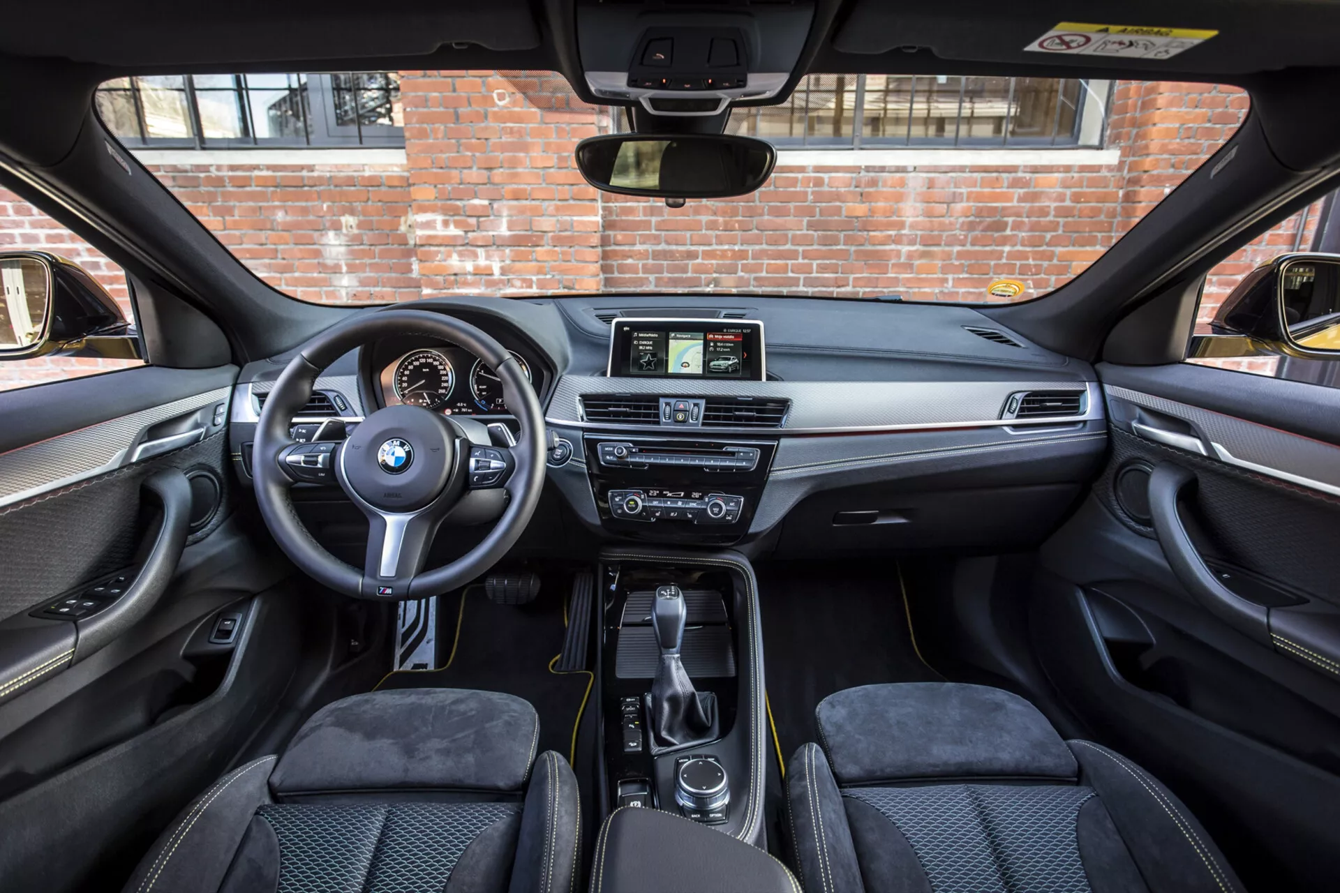 Vue intérieure d'une BMW X2 de 2018, montrant la cabine avant depuis l'arrière des sièges et mettant en évidence le volant avec commandes montées et logo BMW, tableau de bord numérique-analogique, console centrale avec levier de vitesse et système iDrive, affichage infotainment non tactile, tableau de bord luxueux avec garniture en bois ou métallique, contrôle climatique bi-zone, sièges avant profilés, porte-gobelets pratiques et compartiments de rangement, et panneaux de porte avec fonctionnalités de contrôle, sous un toit solaire éclairé par le soleil.