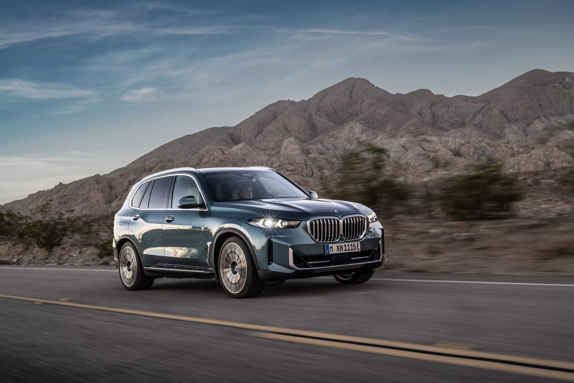 SUV de luxe moderne BMW X5 2023 (G05-F95 Facelift) en mouvement vu depuis un angle de vue avant trois-quarts soulignant le design dynamique, la calandre proéminente en forme de rein, les phares LED élégants, les jantes en alliage à multiples rayons et la silhouette lisse avec peinture métallique