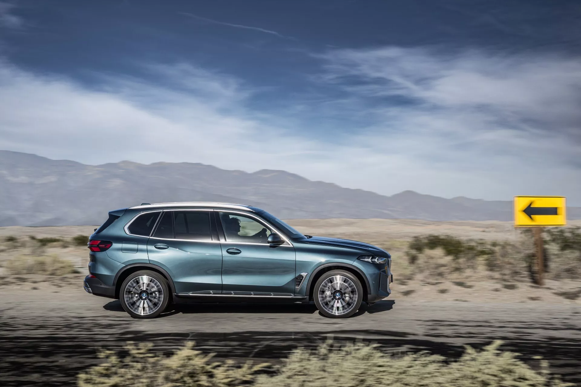 SUV BMW X5 2023 (G05-F95-Facelift) en mouvement, présentant une aérodynamique, des jantes alliage à multi-rayons, des poignées de portes intégrées, des rétroviseurs latéraux aérodynamiques, des vitres teintées, un indicateur latéral, une peinture métallique et un design moderne des feux arrière contre un arrière-plan flou du désert.