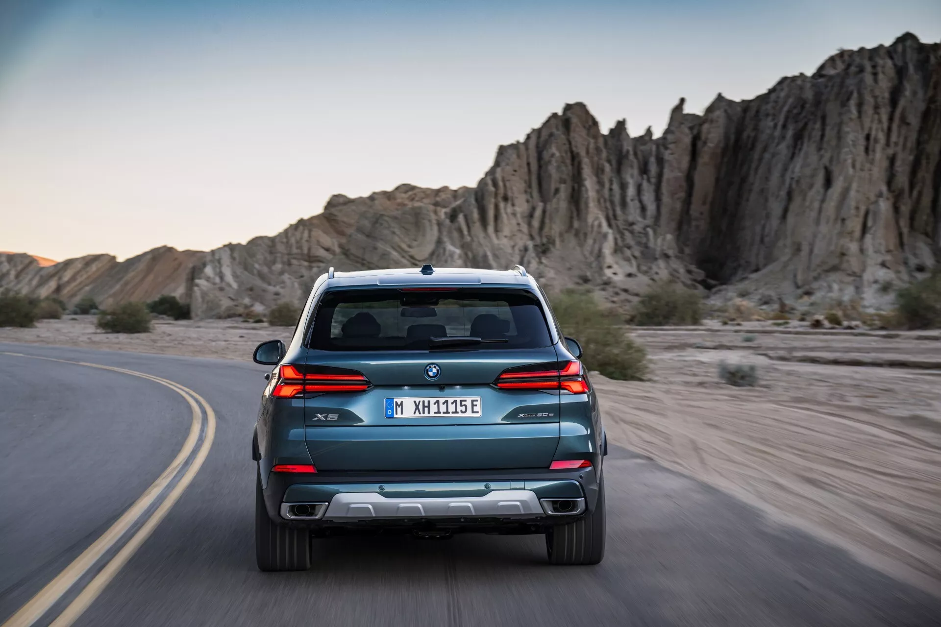 Vue arrière du BMW X5 G05 F95 facelift 2023, circulant sur une route sinueuse, présentant des lignes épurées, un double échappement et des capteurs de stationnement, sur fond de terrain rocheux aventureux