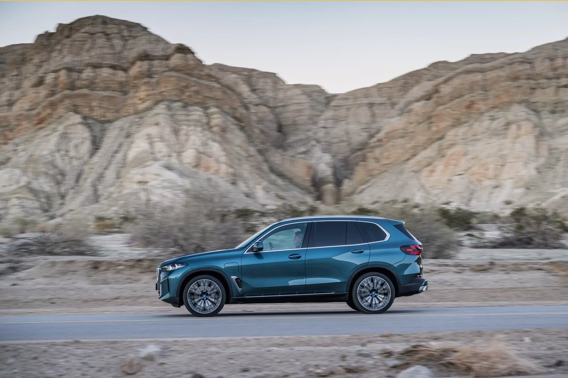 Profil latéral dynamique de la BMW X5 2023 (G05-F95 facelift) en mouvement, SUV bleu en forme de goutte sur une route avec un fond de collines arides au cours de l'heure dorée, mettant en avant un design élégant, une efficacité aérodynamique, des jantes alliage à multiples branches, et un éclairage LED