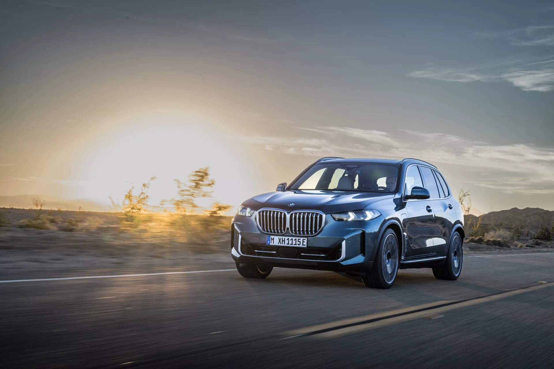 BMW X5 2023 (G05-F95 Facelift) en Mouvement : Vue Dynamique en Plongée Mettant en Avant les Phares LED, Jantes Alliage et Design Aérodynamique sur un Fond de Coucher de Soleil