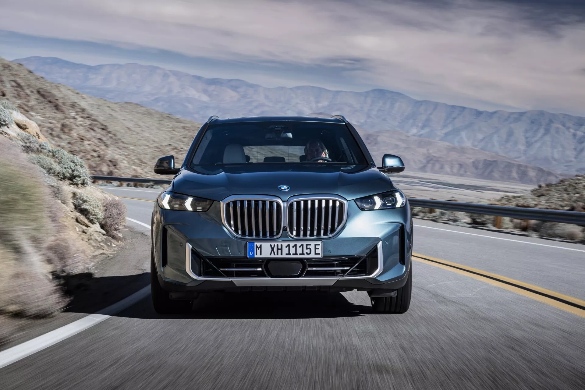 BMW X5 2023 (G05 F95 lifting) en mouvement, capturée sous un angle avant trois quarts mettant en évidence ses phares LED, calandre à naseaux avec volets actifs, roues en alliage et le design aérodynamique élégant sur un fond de désert montagneux, suggérant une technologie avancée et des capacités potentiellement électriques ou hybrides