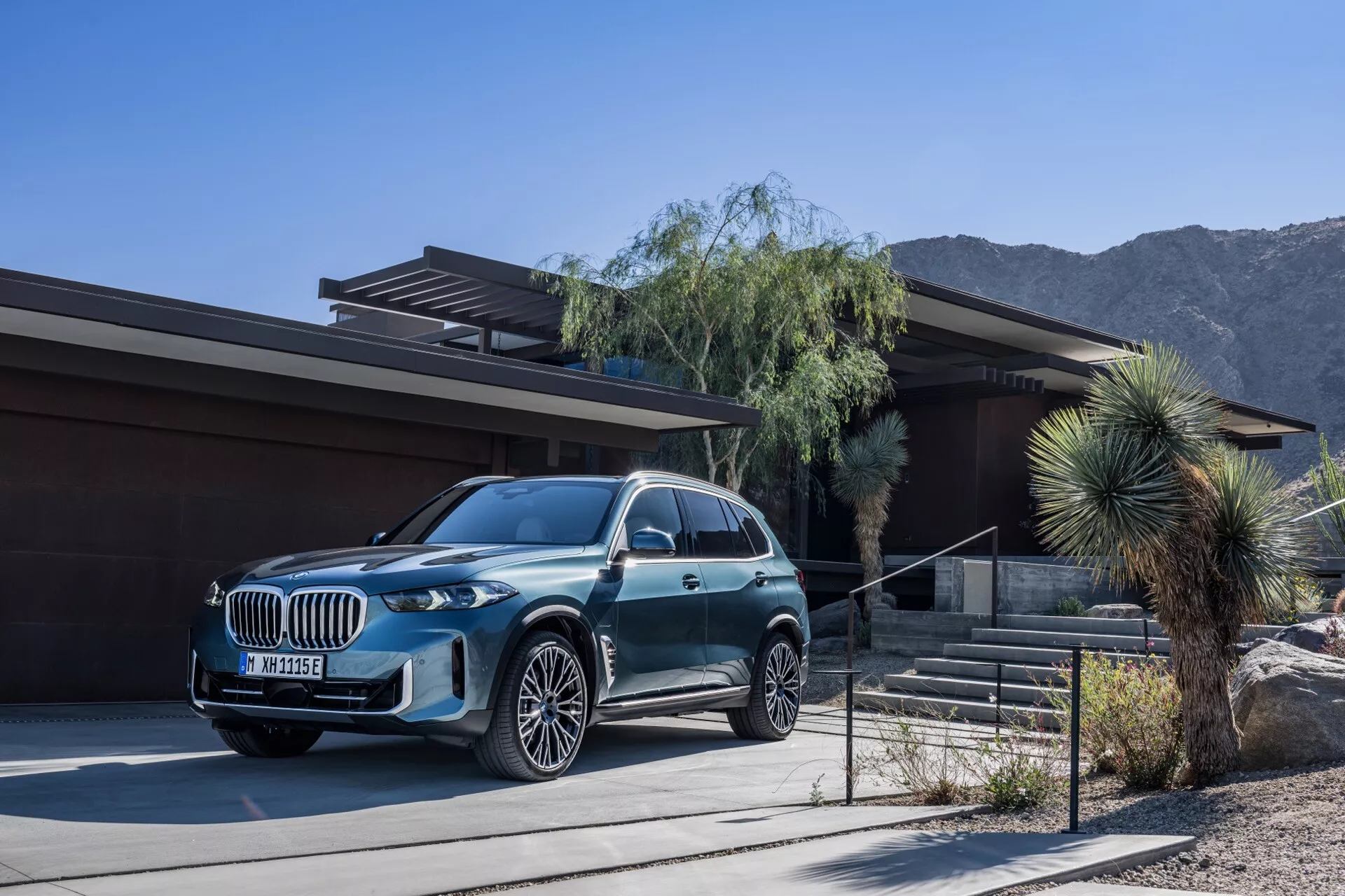SUV de luxe BMW X5 2023 (G05-F95 Facelift) garé devant une maison contemporaine avec un paysage de désert et de montagnes en arrière-plan, présentant une finition métallique, une grande grille, des phares LED et des jantes en alliage à multiples branches