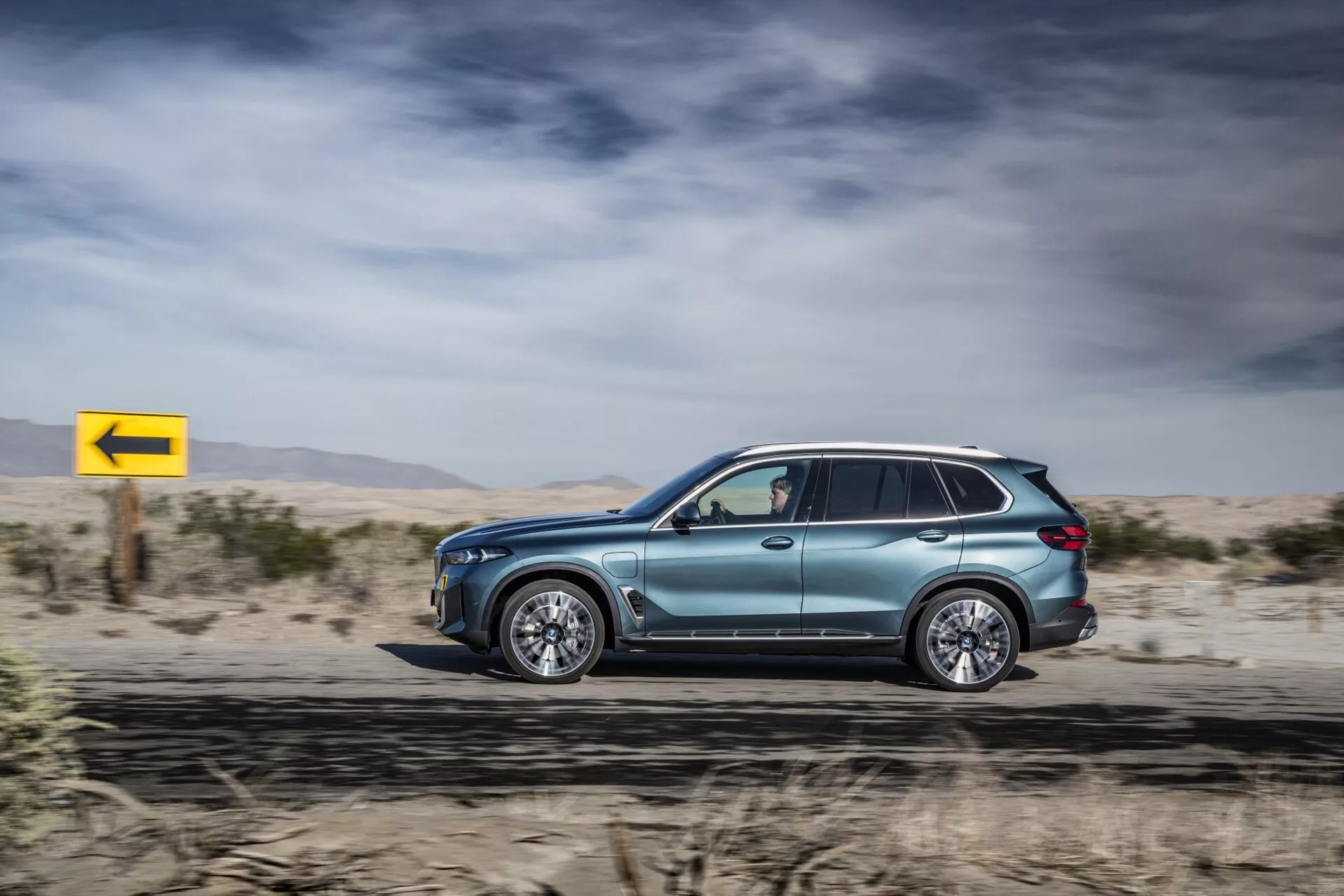 Profil latéral en mouvement du BMW X5 2023 (G05-F95 Facelift), mettant en avant sa peinture métallique, design aérodynamique, jantes en alliage de style luxueux, vitres teintées avec rétroviseurs à clignotants, barres de toit et technologie LED avancée, sur un fond désertique avec un panneau de virage à gauche jaune dynamique.