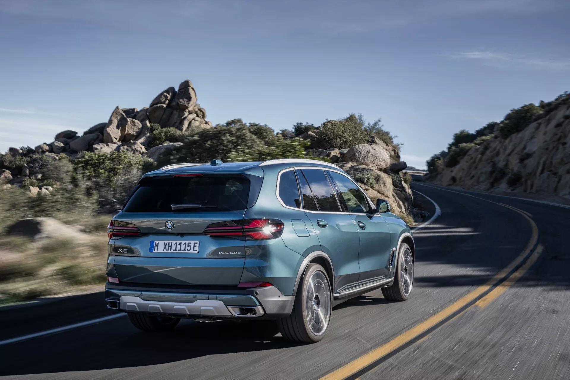 BMW X5 2023 (G05-F95 Facelift) en mouvement, capturée depuis une vue arrière trois quarts mettant en évidence son design SUV sportif, hayon moderne avec feux arrière élégants, jantes alliage bicolores et signes d'électrification sur une route sinueuse sous la lumière du jour claire